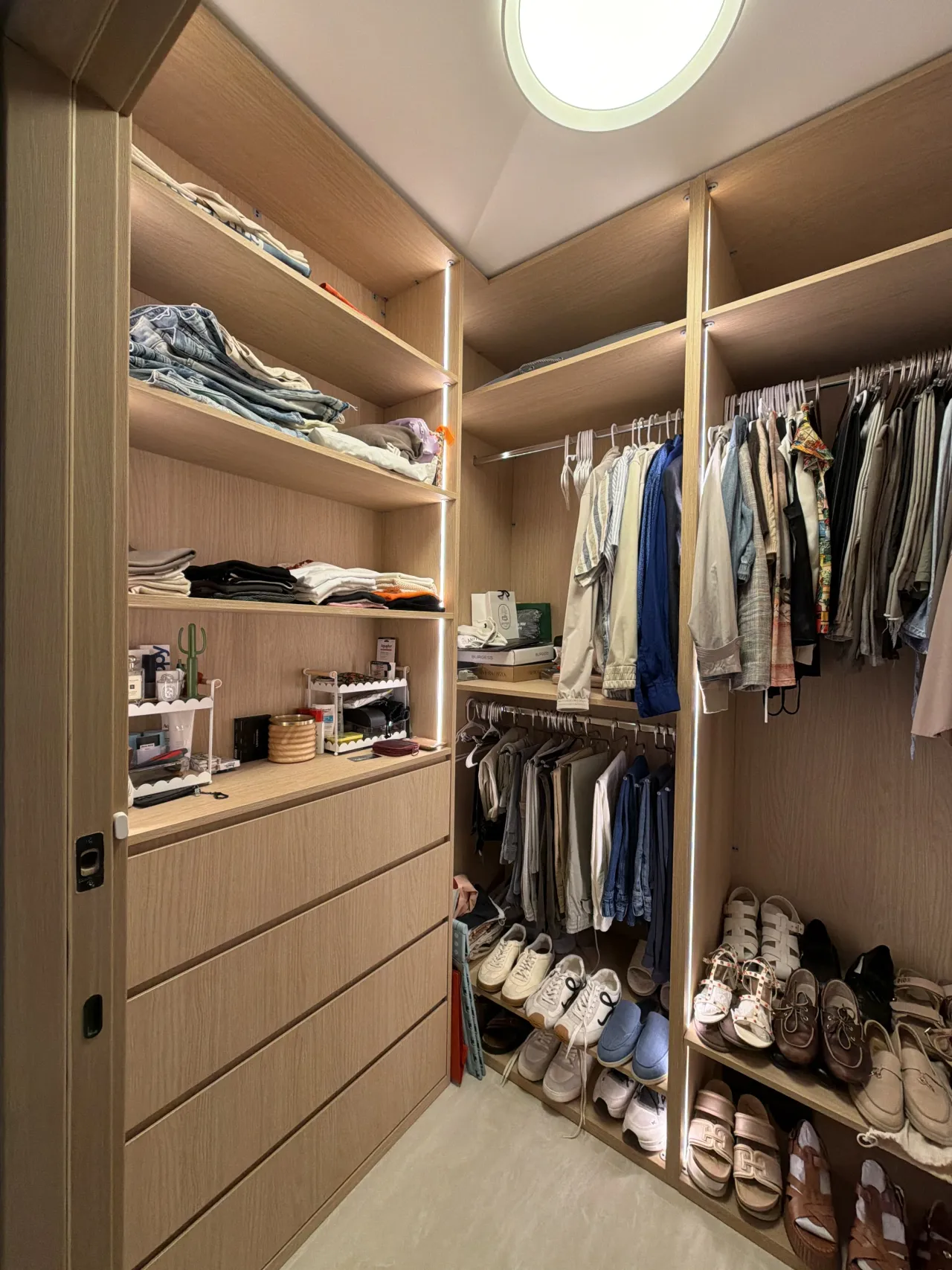 Light wood-grain melamine walk-in closet with shelves of folded clothes, a drawer bank, hanging rods with clothing, shoe shelves and a round ceiling light