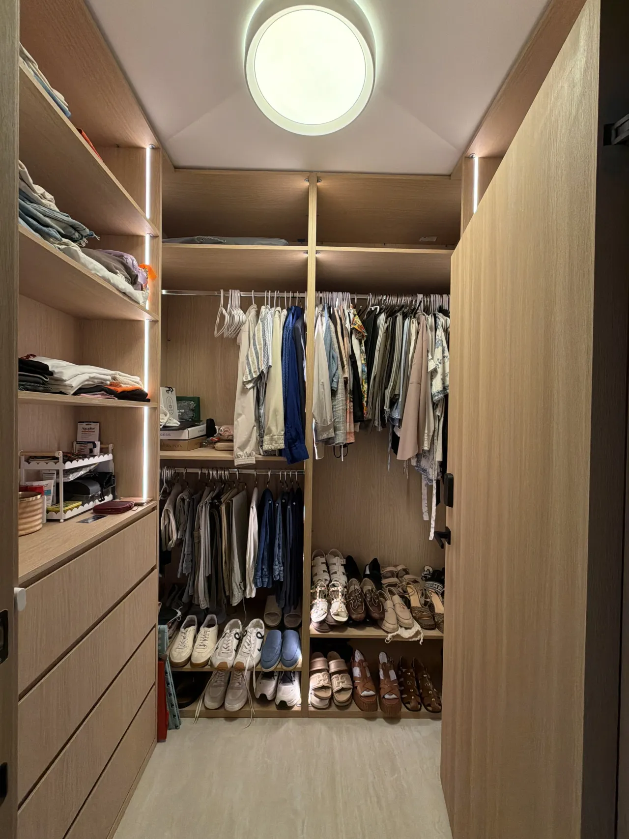 Light wood-grain melamine walk-in closet with a round ceiling light, two tiers of hanging clothes, shoe shelves at the back and a drawer bank at left