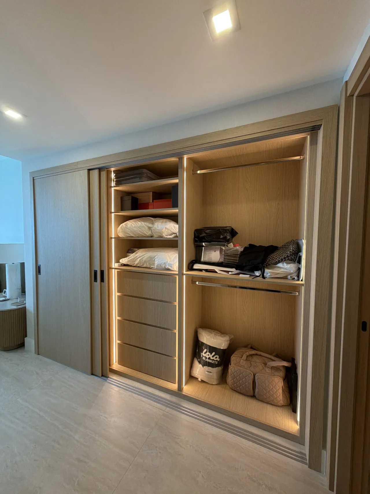 Light wood-grain melamine wardrobe cabinets with sliding doors open showing shelves with folded linens and bags, LED-lit interior, light stone floor