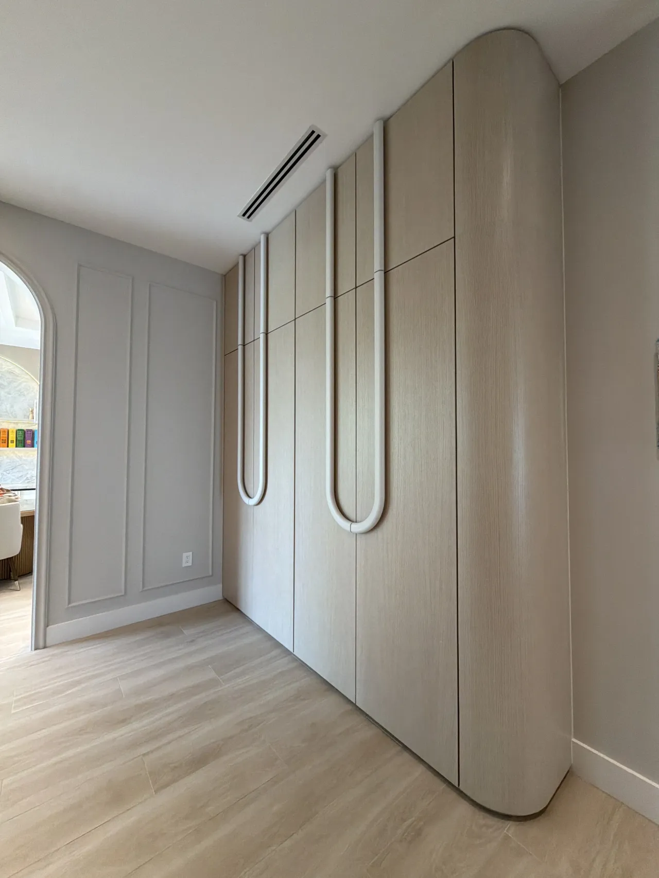 Light wood-grain melamine wardrobe wall with white U-shaped tubular pulls and a rounded corner end panel, next to an open arched opening