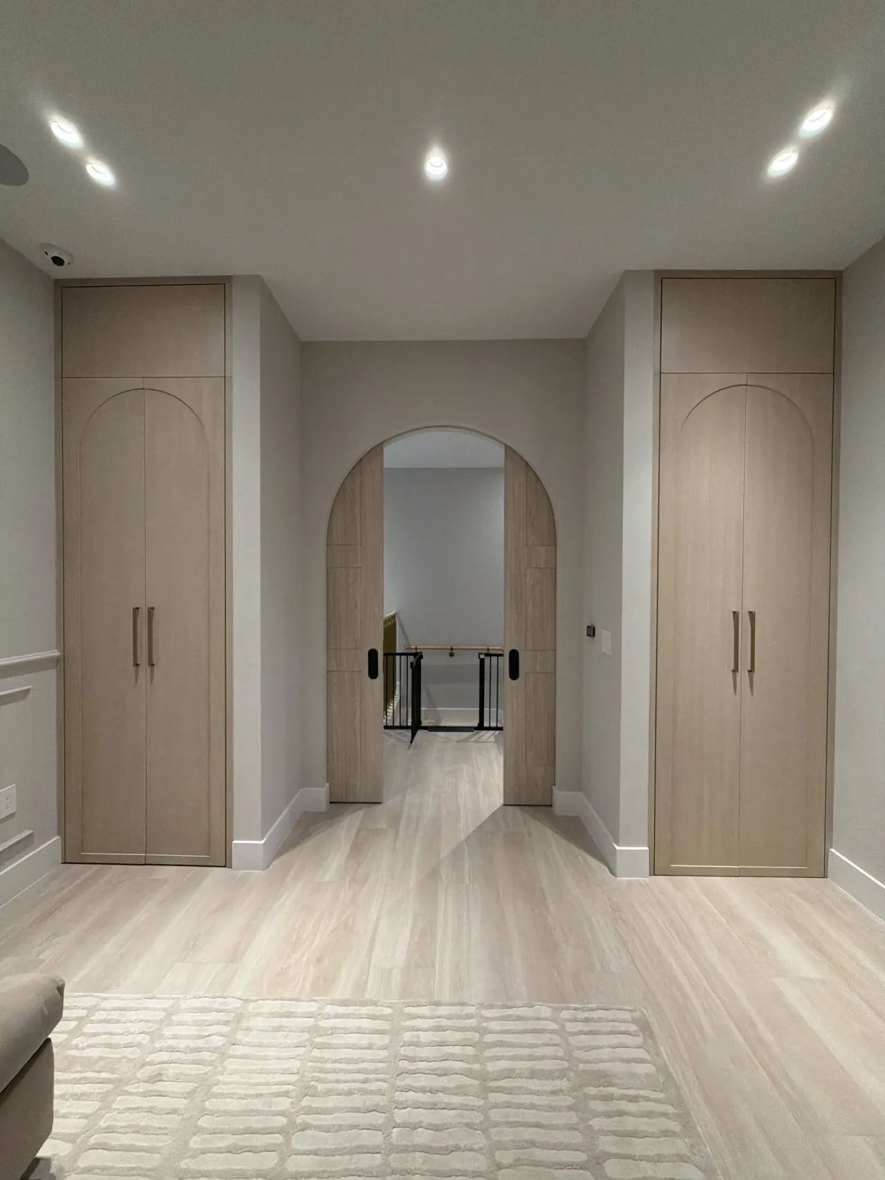 Light wood-grain melamine wardrobe wall with tall arched-panel doors and gold-tone bar handles flanking a hallway with an arched melamine double door