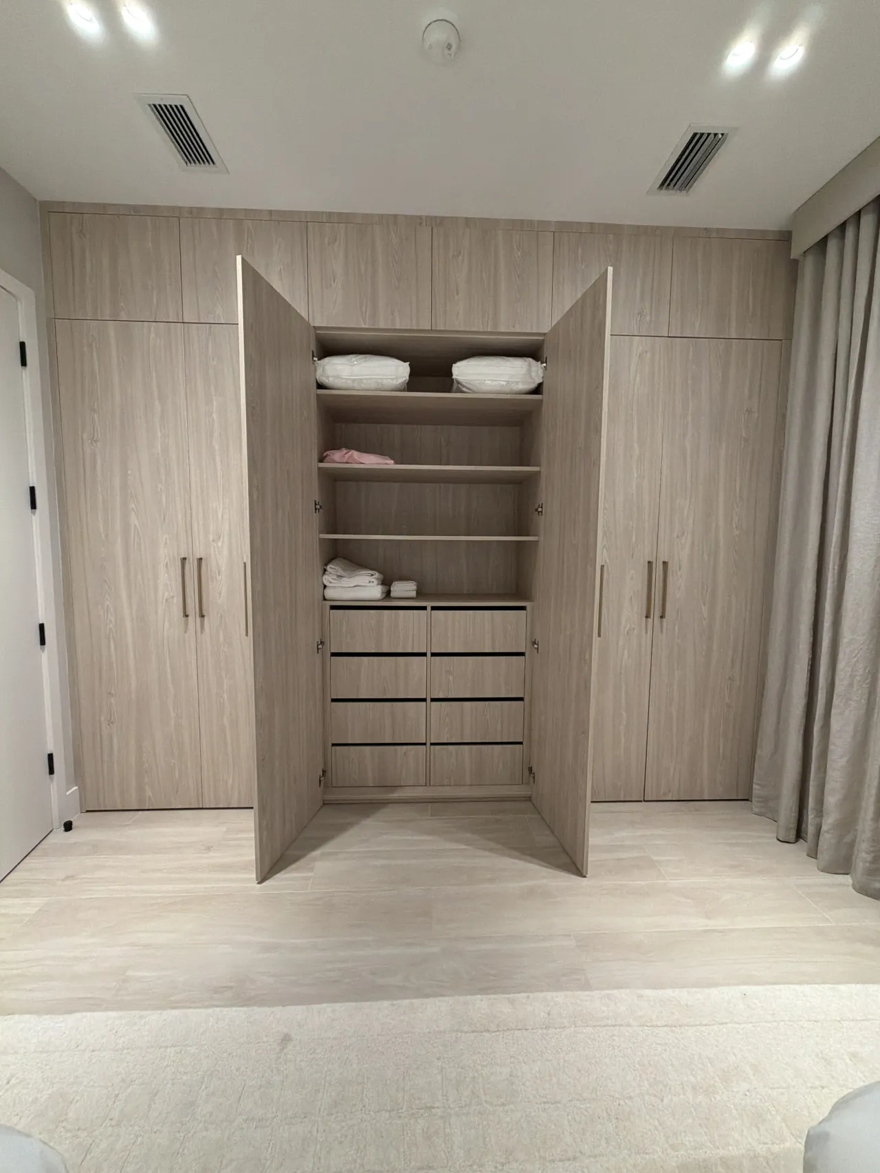 Light wood-grain melamine wardrobe wall with a center pair of doors open, showing shelves of folded linens and a bank of drawers