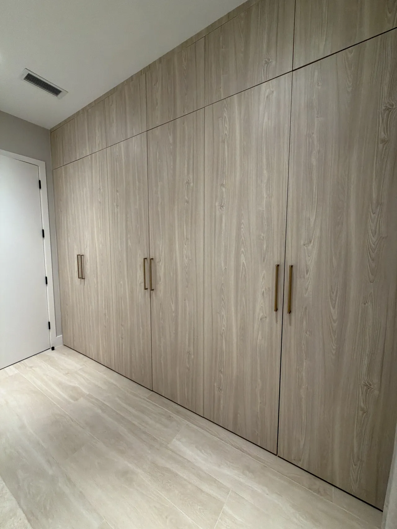 Light wood-grain melamine wardrobe wall with slim gold-tone bar handles, beside a white door on light stone-look tile