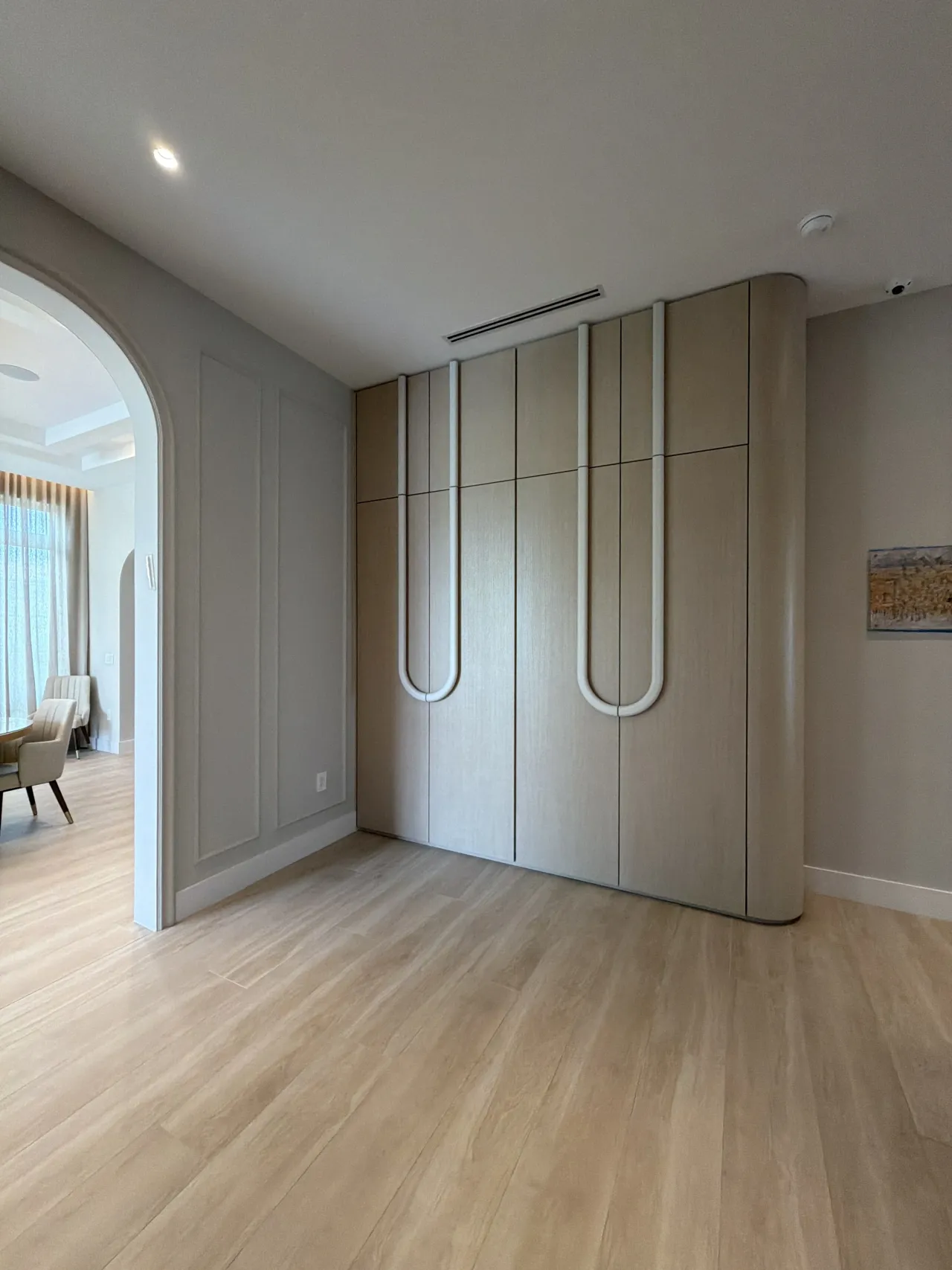 Light wood-grain melamine wardrobe with white U-shaped tubular pulls and a rounded end, beside an open arched opening to a room with a chair