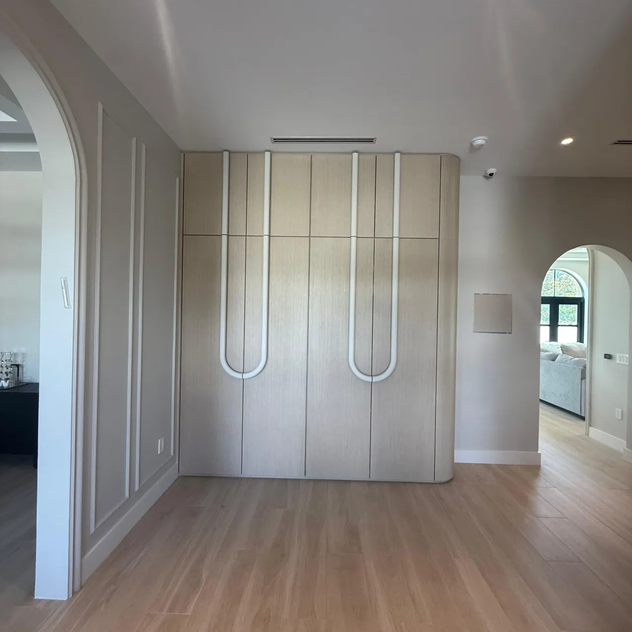 Light wood wardrobe wall with curved white U-shaped pull handles beside an arched doorway leading to a living room, light wood floor