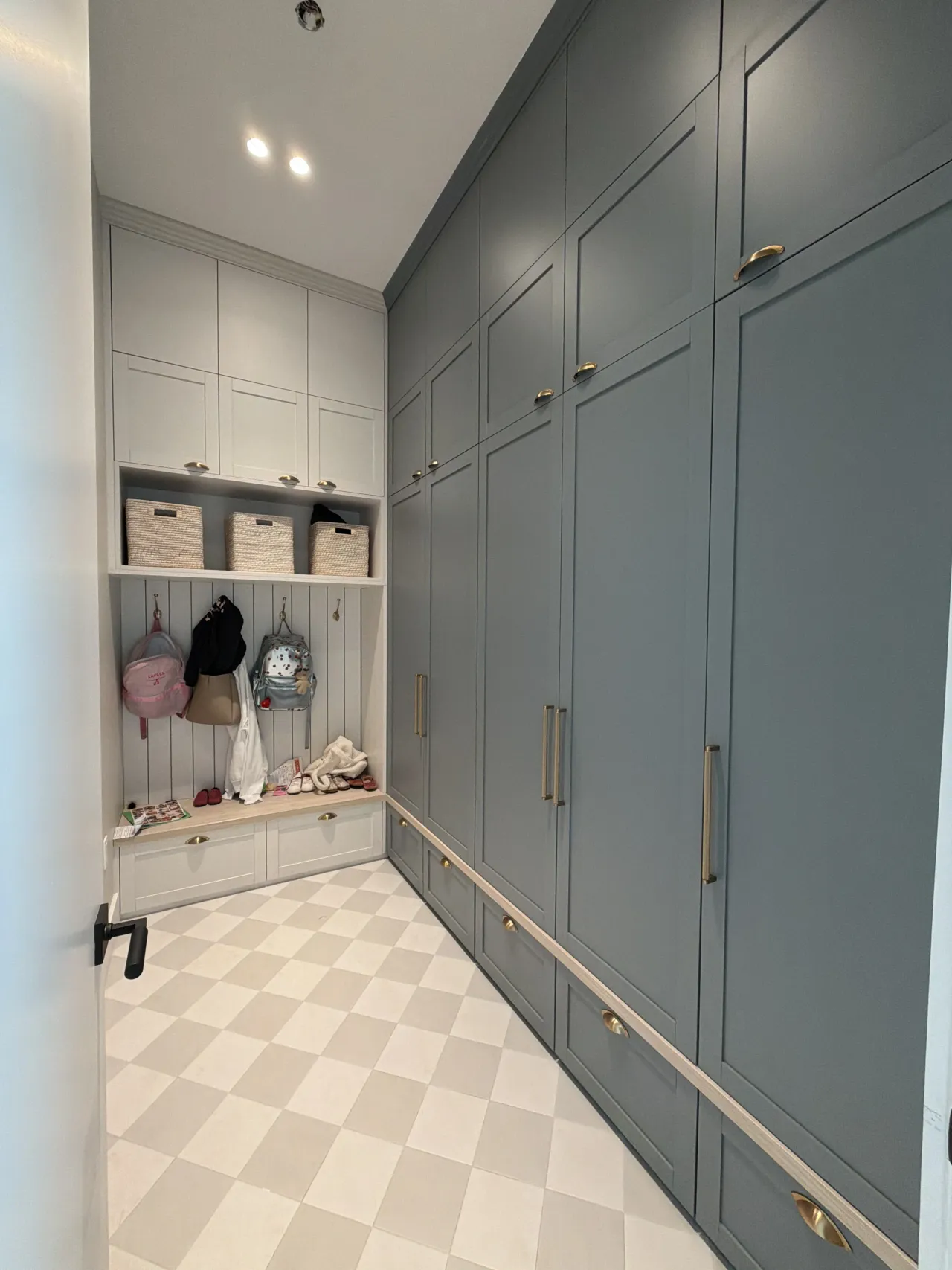 Mudroom-style closet with blue-gray shaker-style cabinet doors, gold-tone hardware, an open cubby with baskets, a bench, and a harlequin-pattern tile floor
