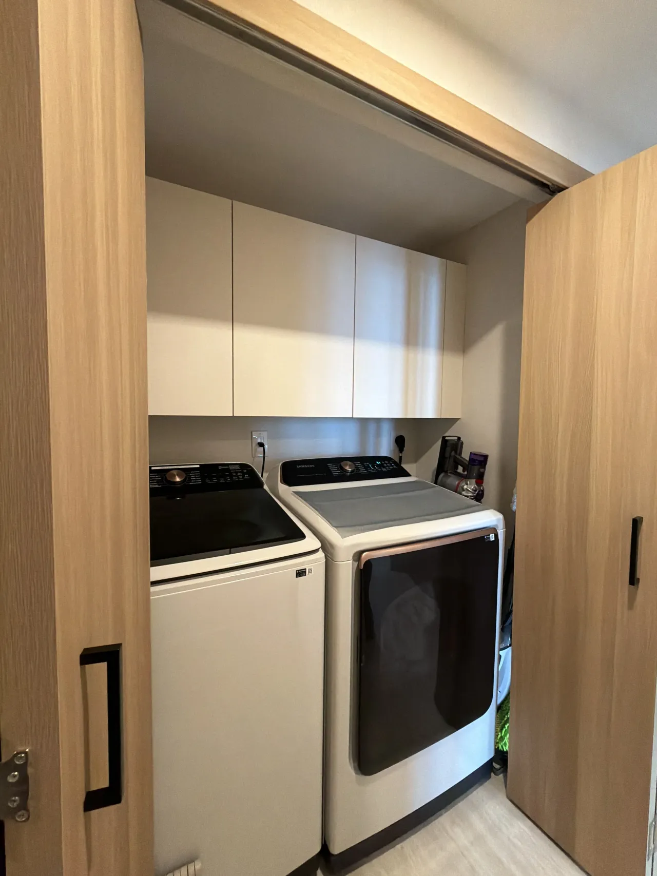 Open laundry closet with light wood-grain melamine doors showing a washer and dryer beneath beige upper cabinets, second closet visible