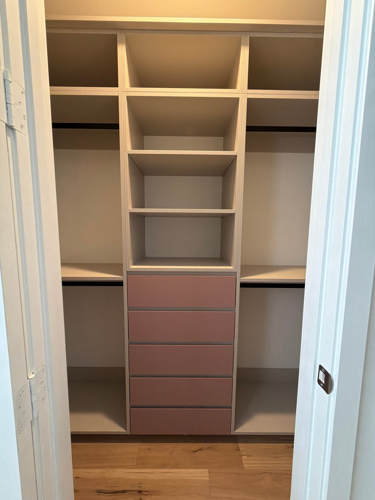 Reach-in closet with cream shelving, black hanging rods on both sides and a centre stack of five mauve-pink drawers above a wood-grain floor