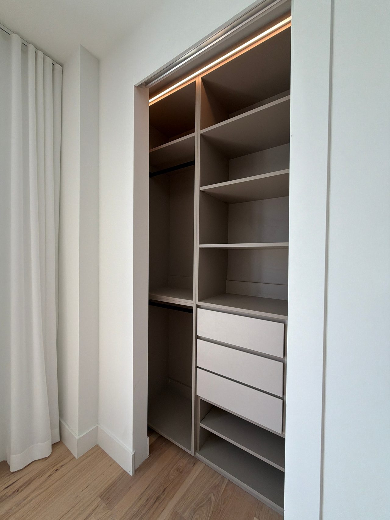 Reach-in closet with greige open shelving, two hanging rods, a stack of white drawer fronts and an LED strip along the top edge