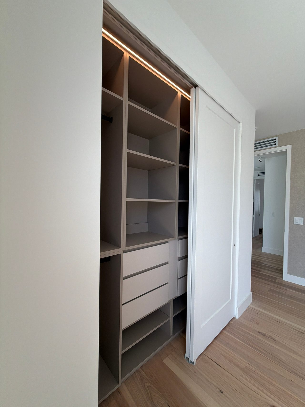 Reach-in closet with a white sliding door pushed aside, showing greige open shelving, hanging rods, white drawer fronts and an LED strip