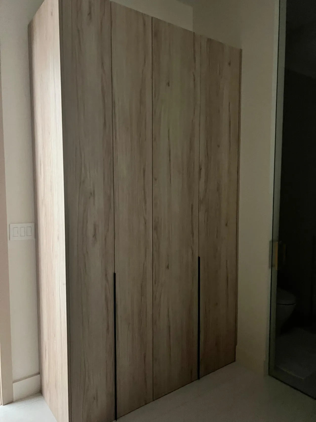 Rustic light wood wardrobe with four doors and black vertical handles, dim lighting, near a glass door leading to a bathroom
