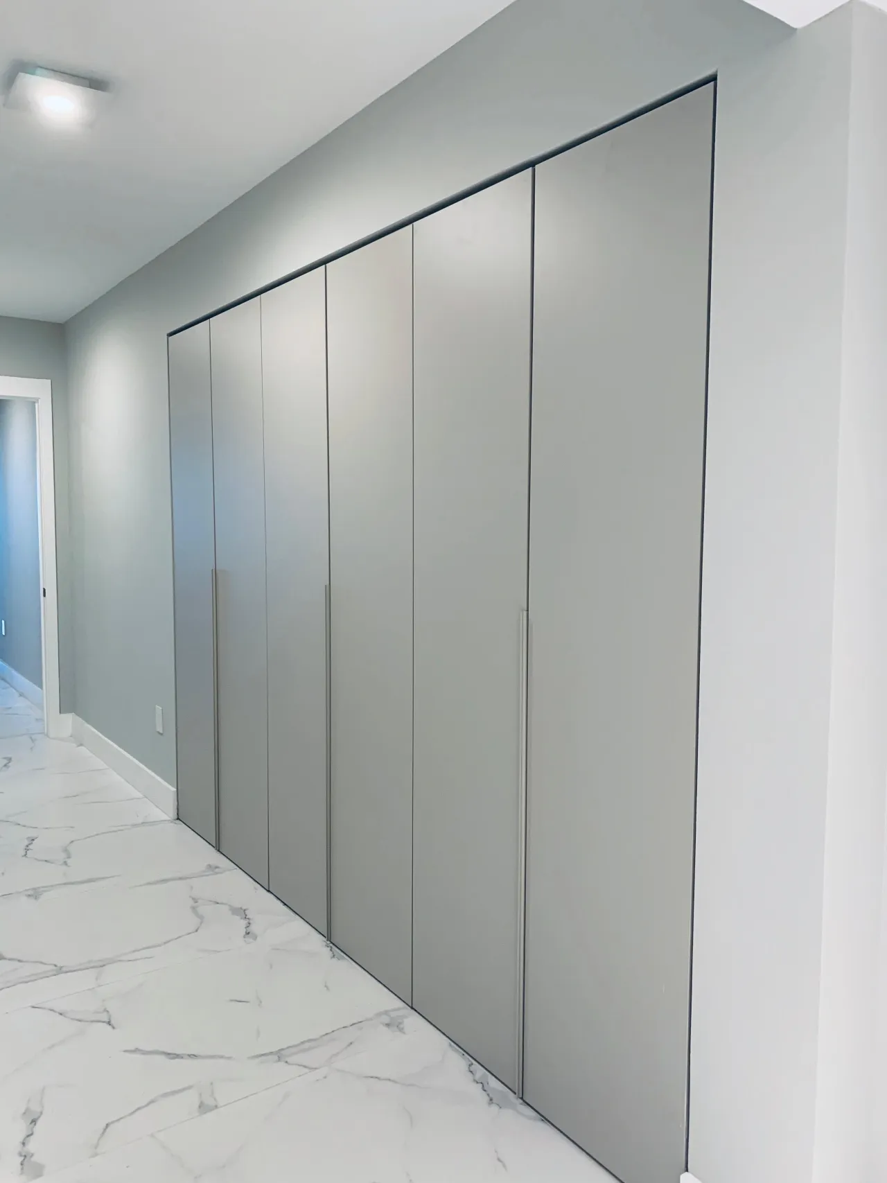 Sage gray floor-to-ceiling wardrobe with thin vertical handles along a veined stone hallway, angled perspective toward a doorway at the end