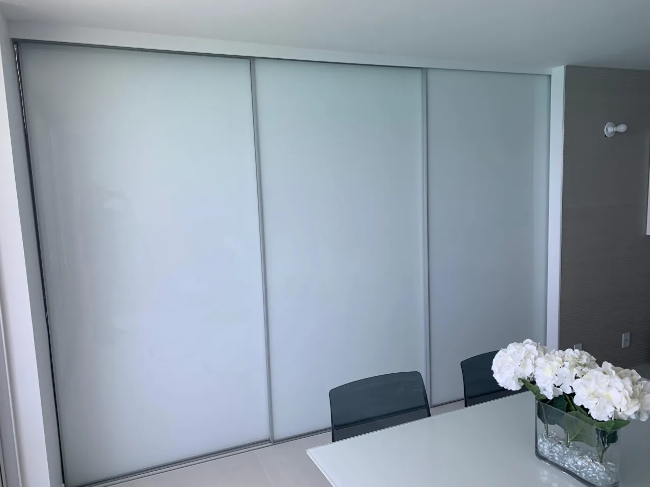 Three sliding closet doors in frosted white glass with slim metal frames and a top track, beside a dark textured wall panel, white table in front