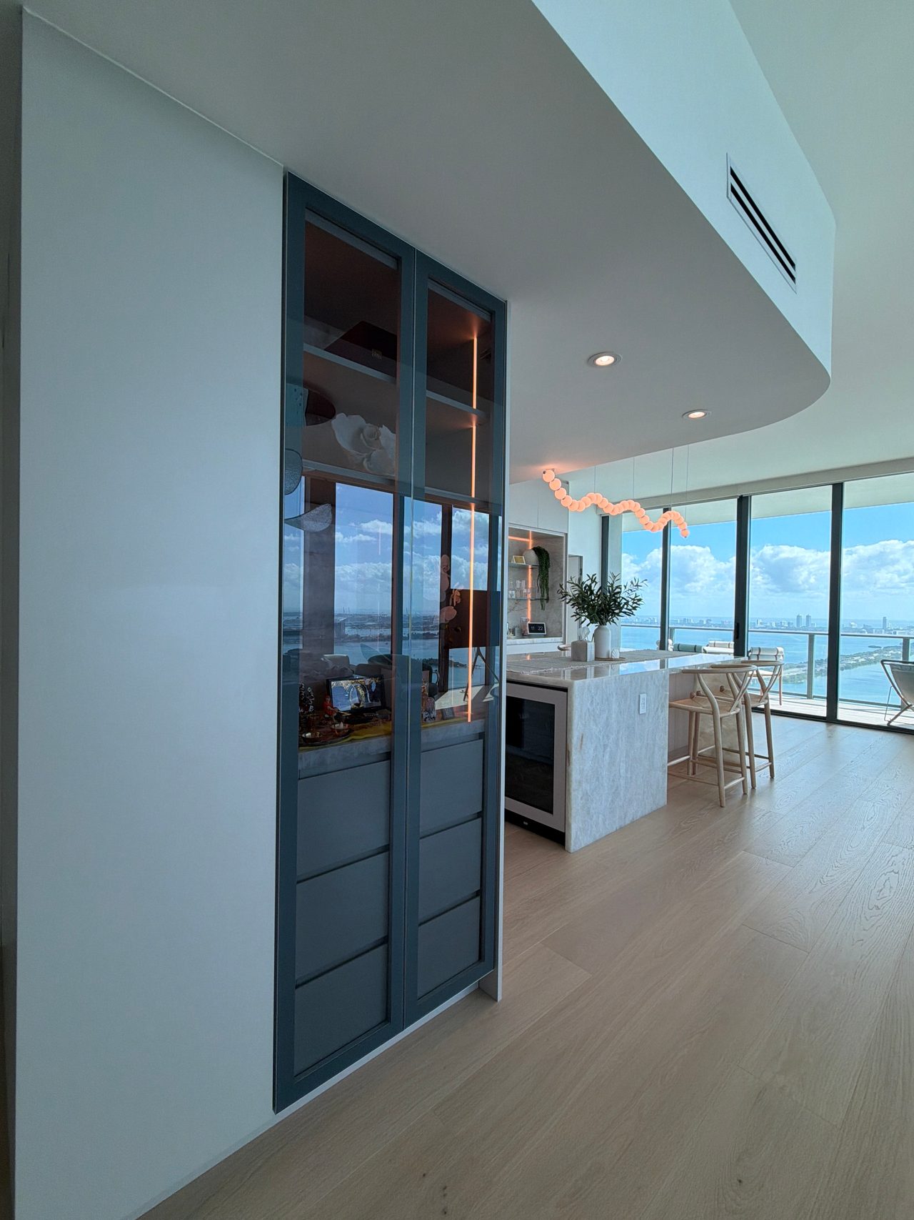 Tall built-in cabinet with dark blue-grey framed glass doors over drawers, opening to a kitchen island and floor-to-ceiling windows over water