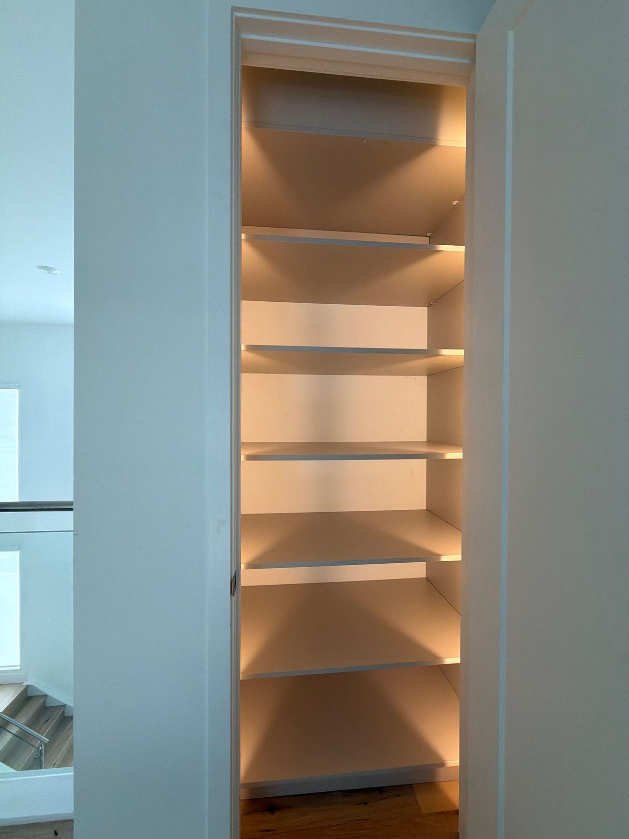 Tall built-in niche with six white shelves and warm LED strip lighting, set into a white wall beside a stair landing