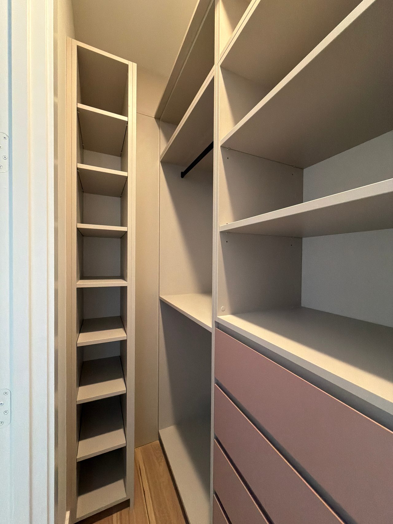 Narrow closet with a tall cubby column at left, cream open shelving with a black hanging rod, and mauve-pink drawer fronts at right