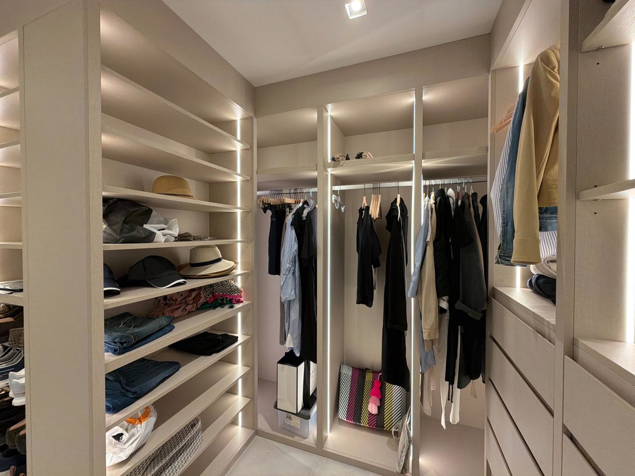 Walk-in closet with a tall gray melamine shelving stack lit by LED strips, two full-height hanging bays and a drawer bank opposite