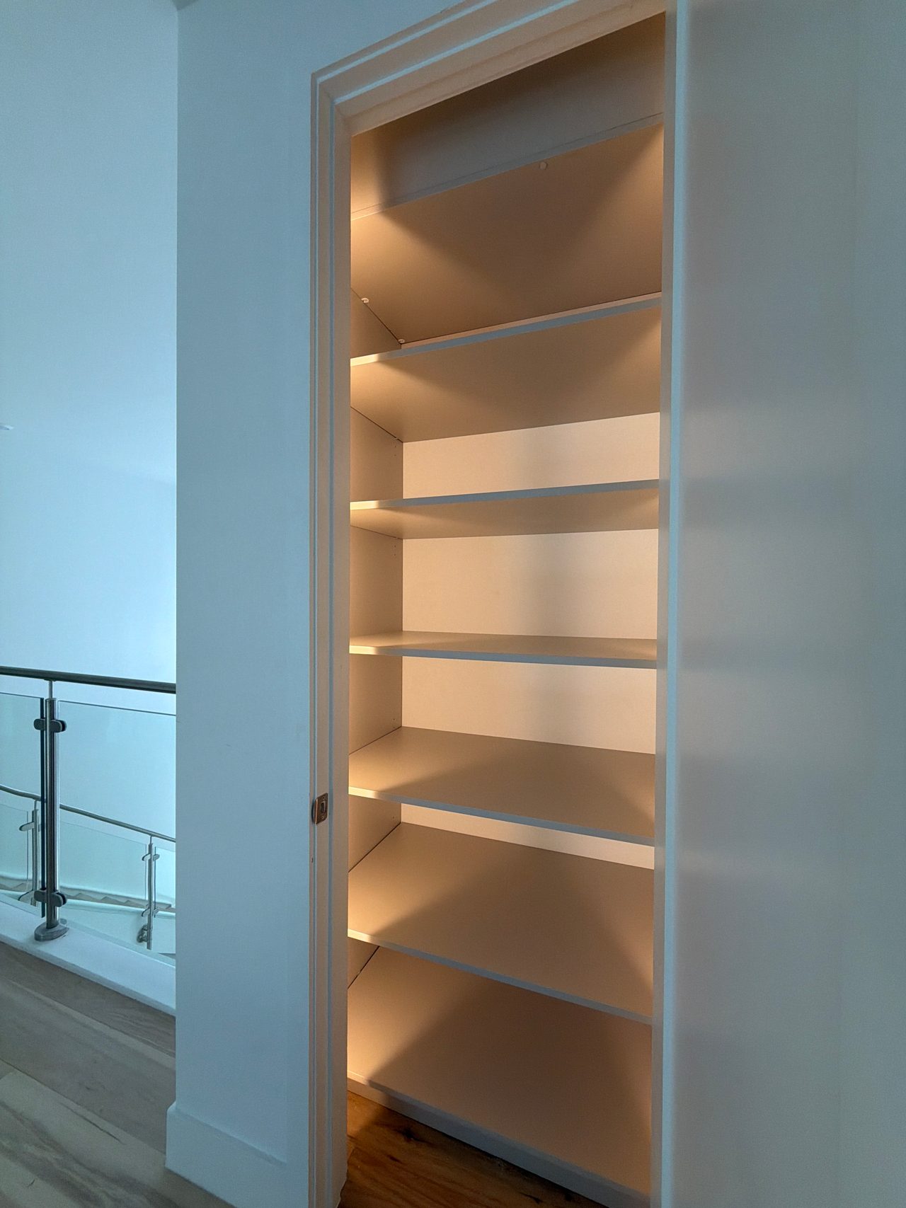 Tall recessed shelving niche with six white shelves lit by warm LED strips, next to a glass-panel railing and a wood-grain floor