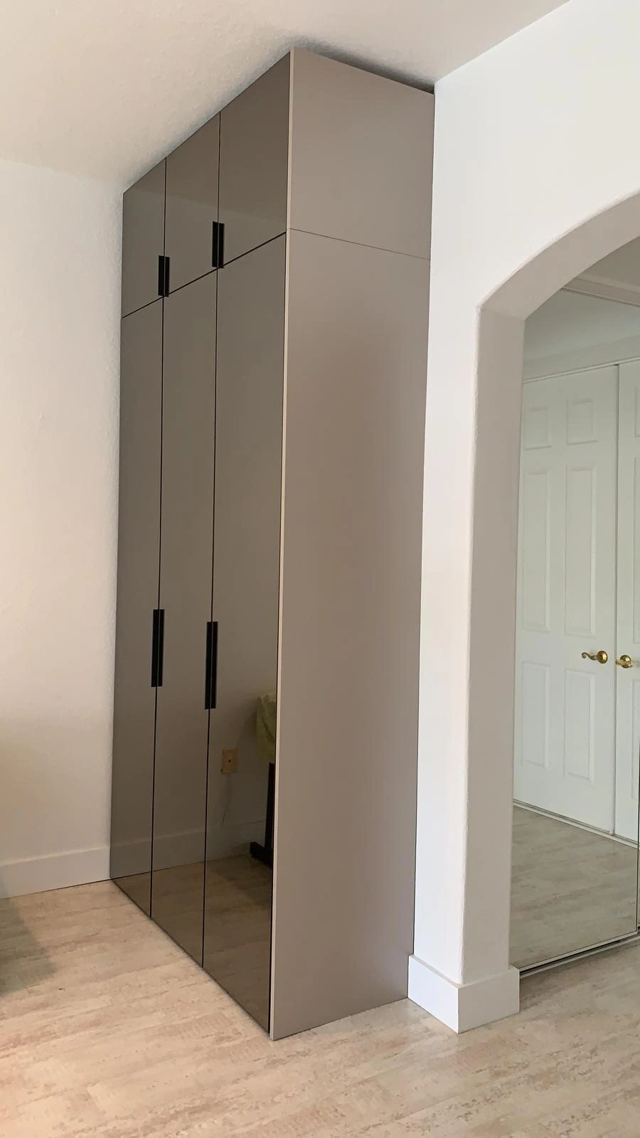 Tall taupe wardrobe with tinted mirror door fronts and slim black vertical handles, beside an arched opening, light wood-look floor