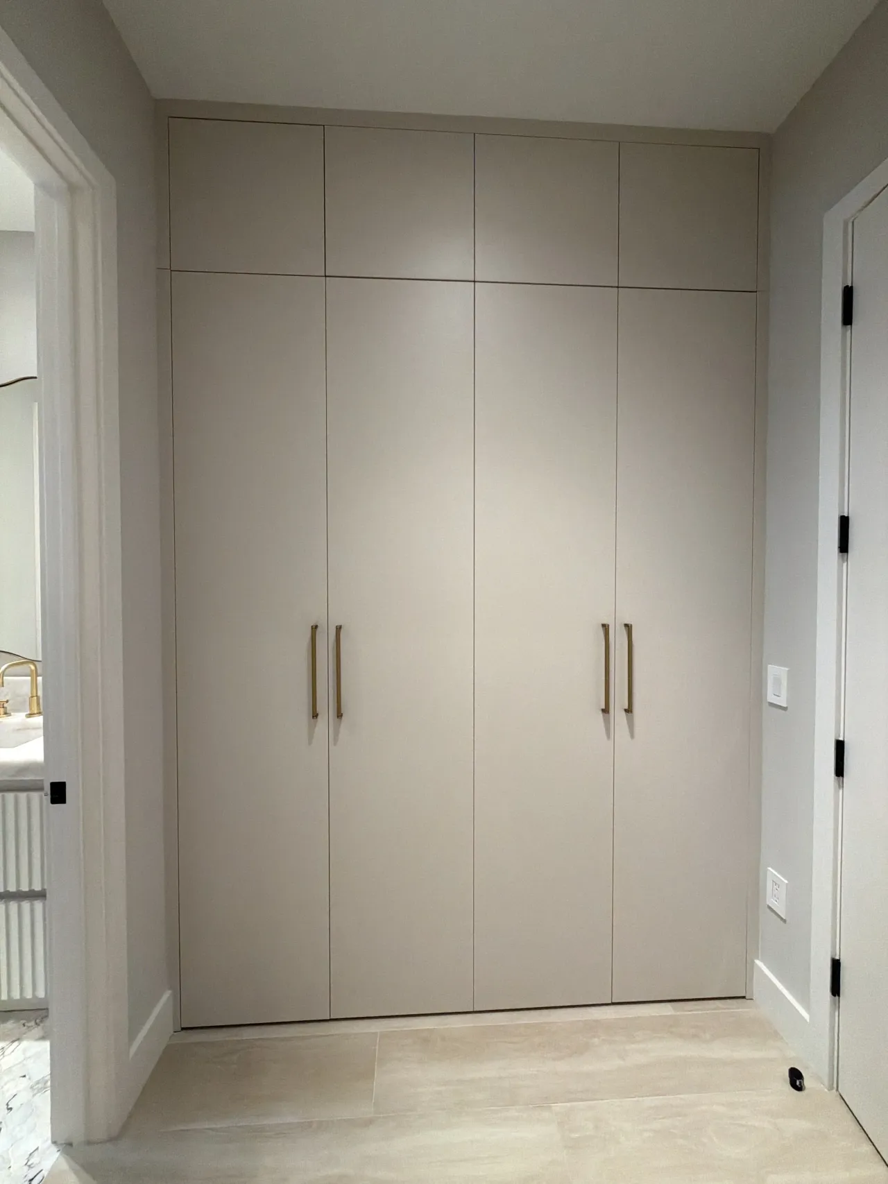 Taupe floor-to-ceiling wardrobe with gold-tone bar pulls in an alcove, beside an open doorway showing a vanity with a gold-tone faucet