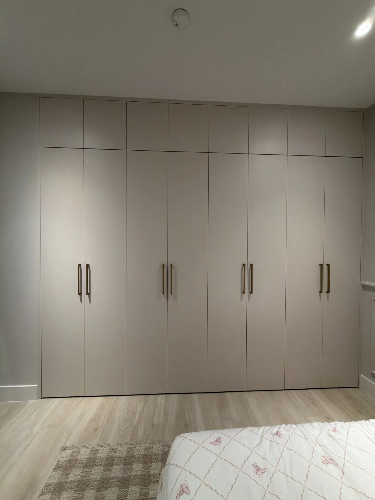 Taupe floor-to-ceiling wardrobe wall with slim gold-tone bar pulls, a checked rug on the wood-look floor and a bed with a pink-patterned cover