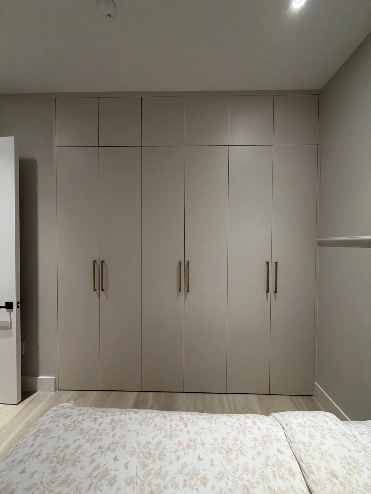 Taupe floor-to-ceiling wardrobe wall with upper cabinets and slim gold-tone bar pulls, seen across a bed with a cream floral cover