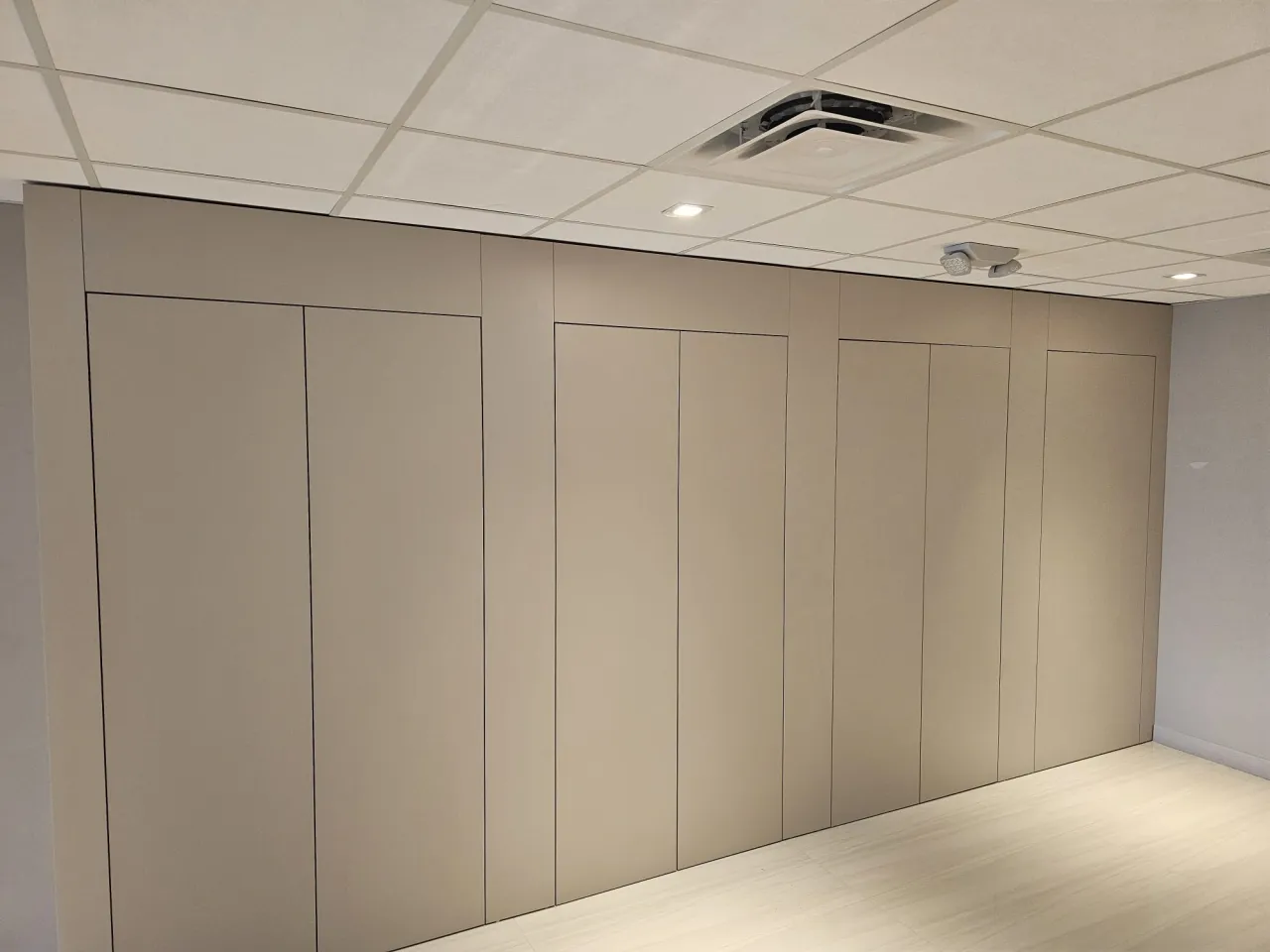 Taupe full-height cabinet wall with recessed panel doors running the length of a room, suspended grid tile ceiling with a vent and downlights