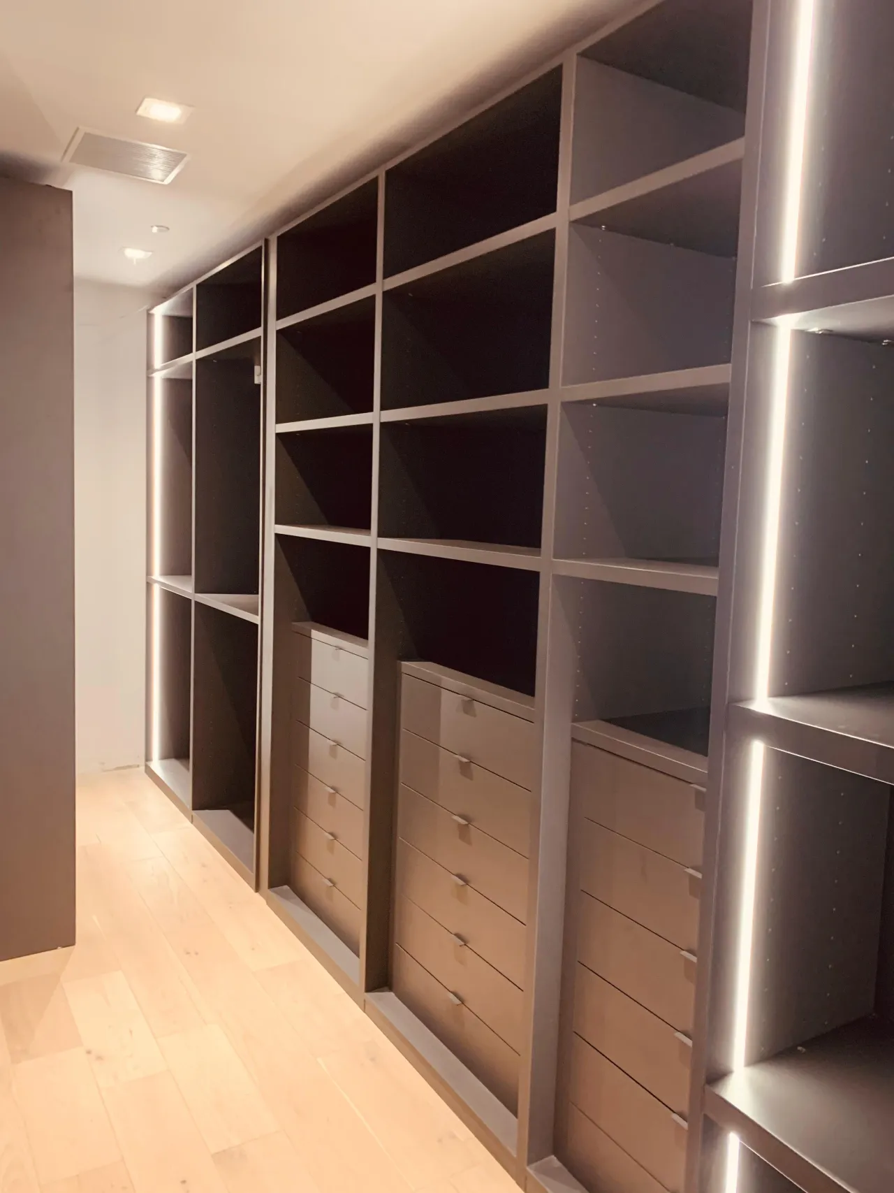 Taupe-gray open cubby shelving wall with drawer stacks below and vertical LED strip lighting, light wood plank floor in a narrow room