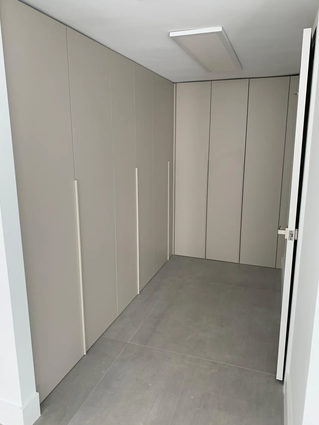Taupe L-shaped floor-to-ceiling wardrobe wall with long vertical bar handles, a flush ceiling light panel and large-format gray tile floor