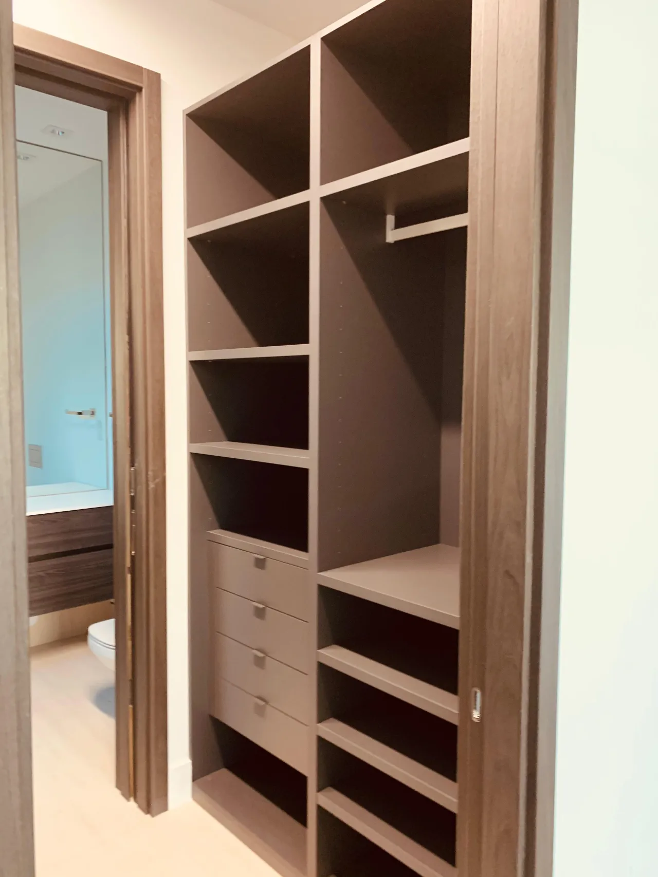 Small closet interior in warm taupe with open shelving, a hanging rod, and a four-drawer stack with small round pulls, dark wood door casing at left