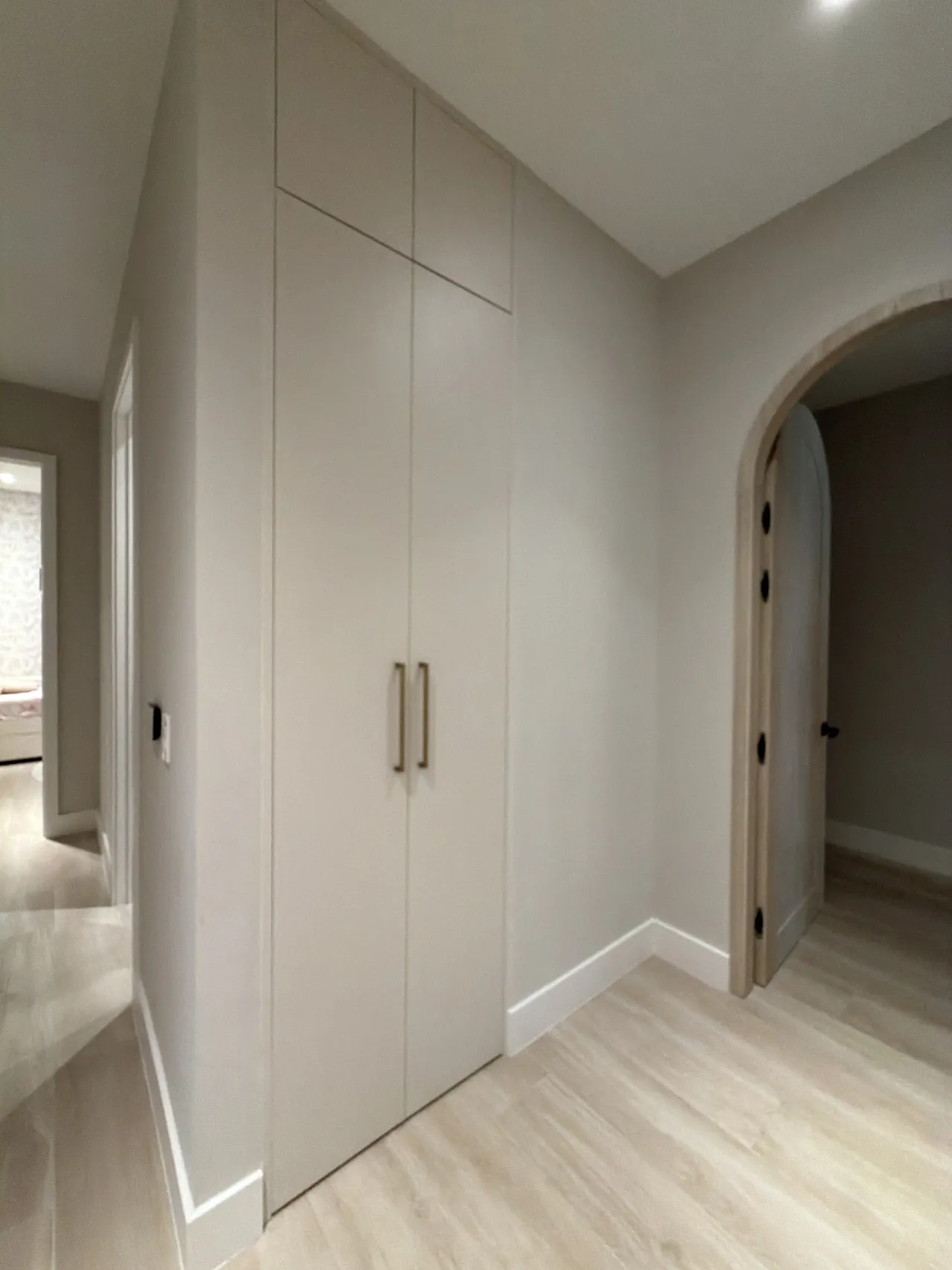 Taupe wardrobe with gold-tone bar pulls in a hallway corner, beside an arched doorway with a light wood-frame arched door on wood-look floor