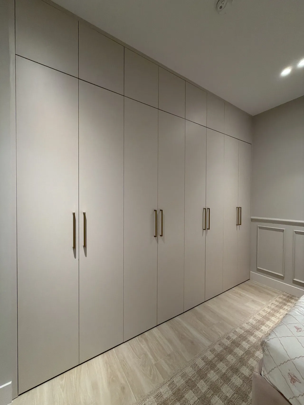 Taupe wardrobe wall of flat-panel doors with gold-tone bar pulls, beside a bed with a checked rug on light wood-look flooring