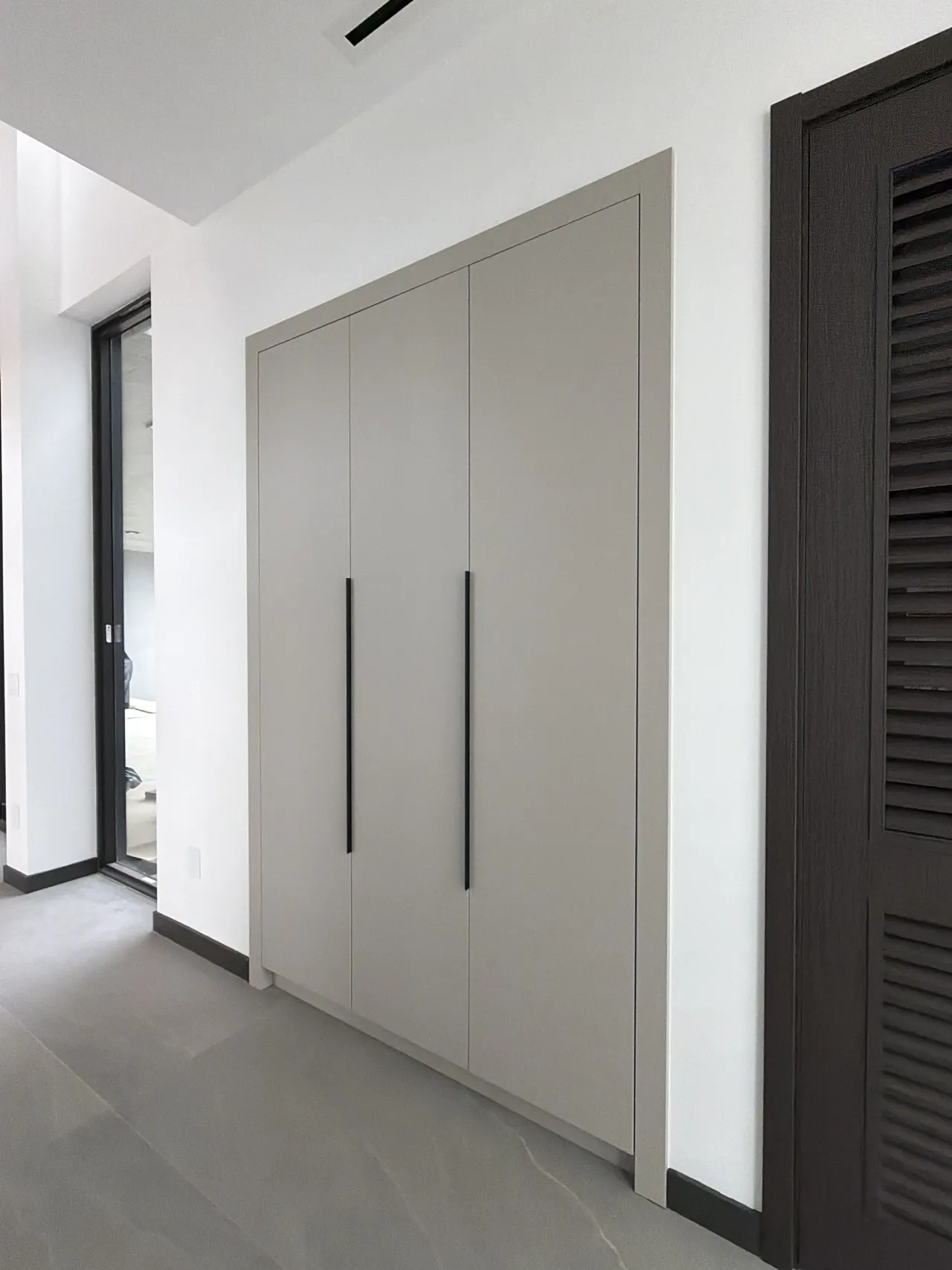 Three-door gray wardrobe with black vertical pulls in a hallway beside a louvered dark wood door and a glass-front doorway
