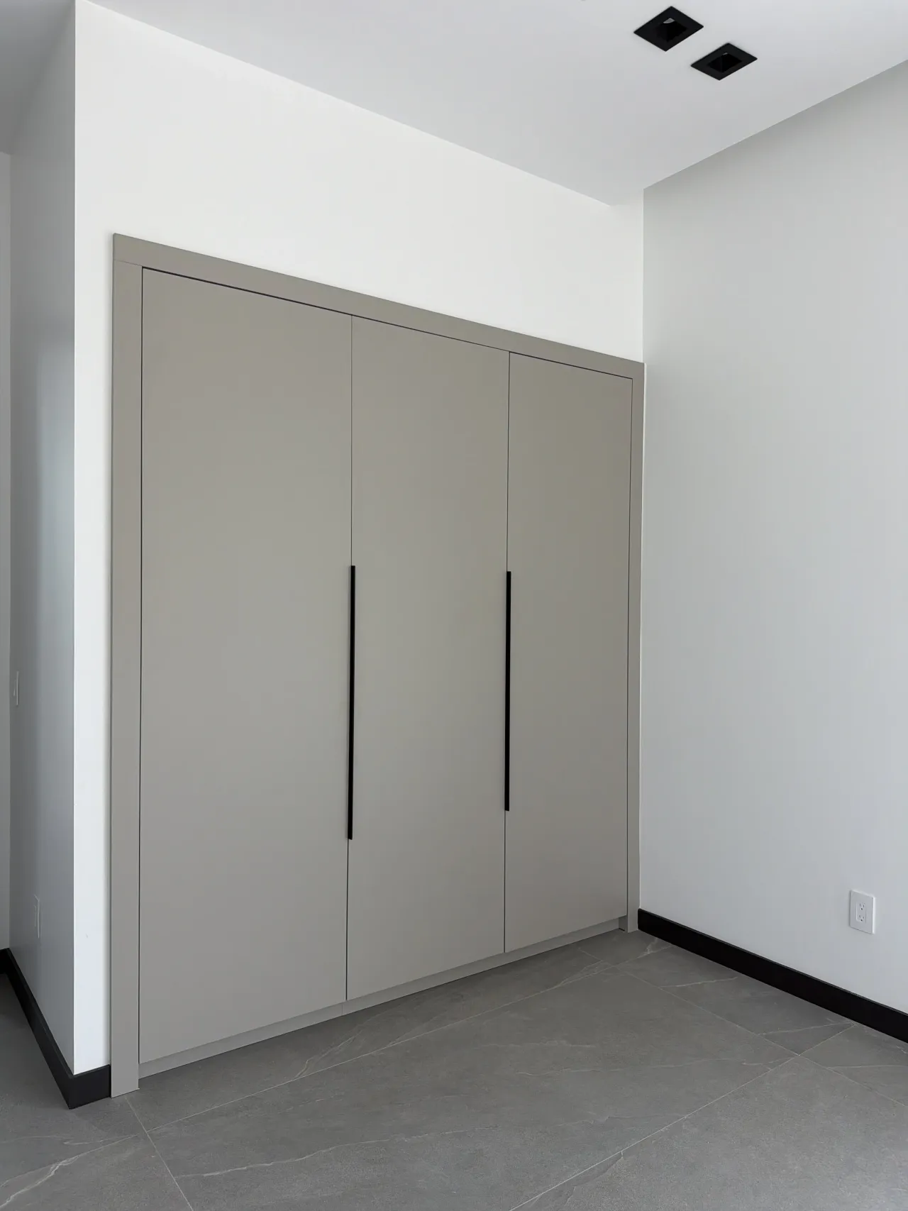 Three-door gray wardrobe in a corner nook with black vertical pulls and a gray stone tile floor