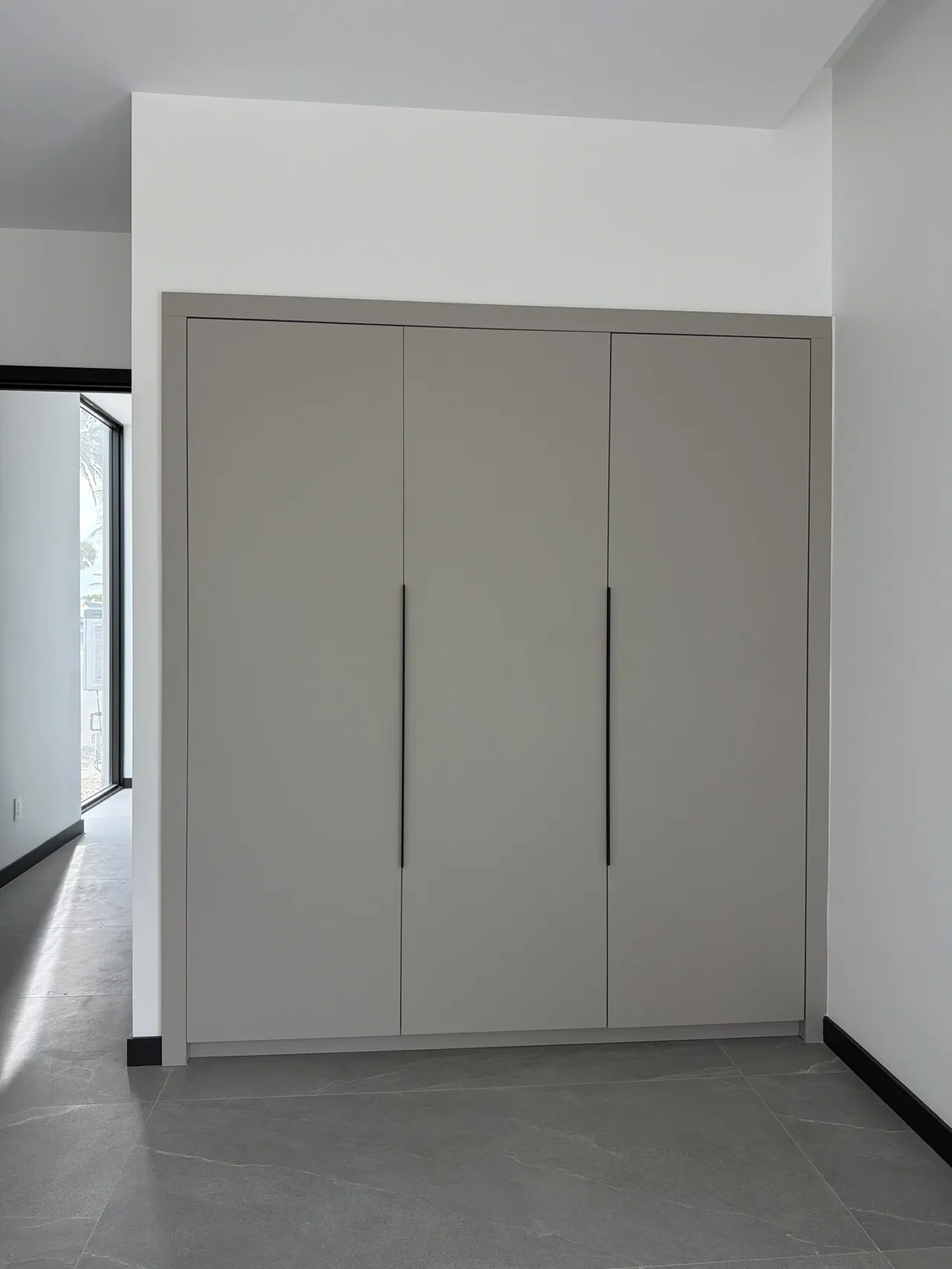 Three-door greige wardrobe with black vertical pulls set into a wall niche, beside a corridor with floor-to-ceiling glazing and dark gray tile