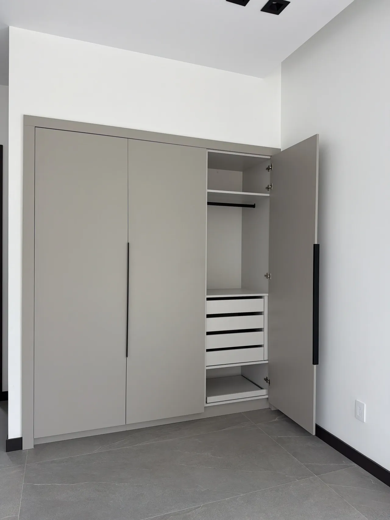 Three-door greige wardrobe with the right door open, showing a white interior with shelf, hanging rod and four white drawers, gray tile floor