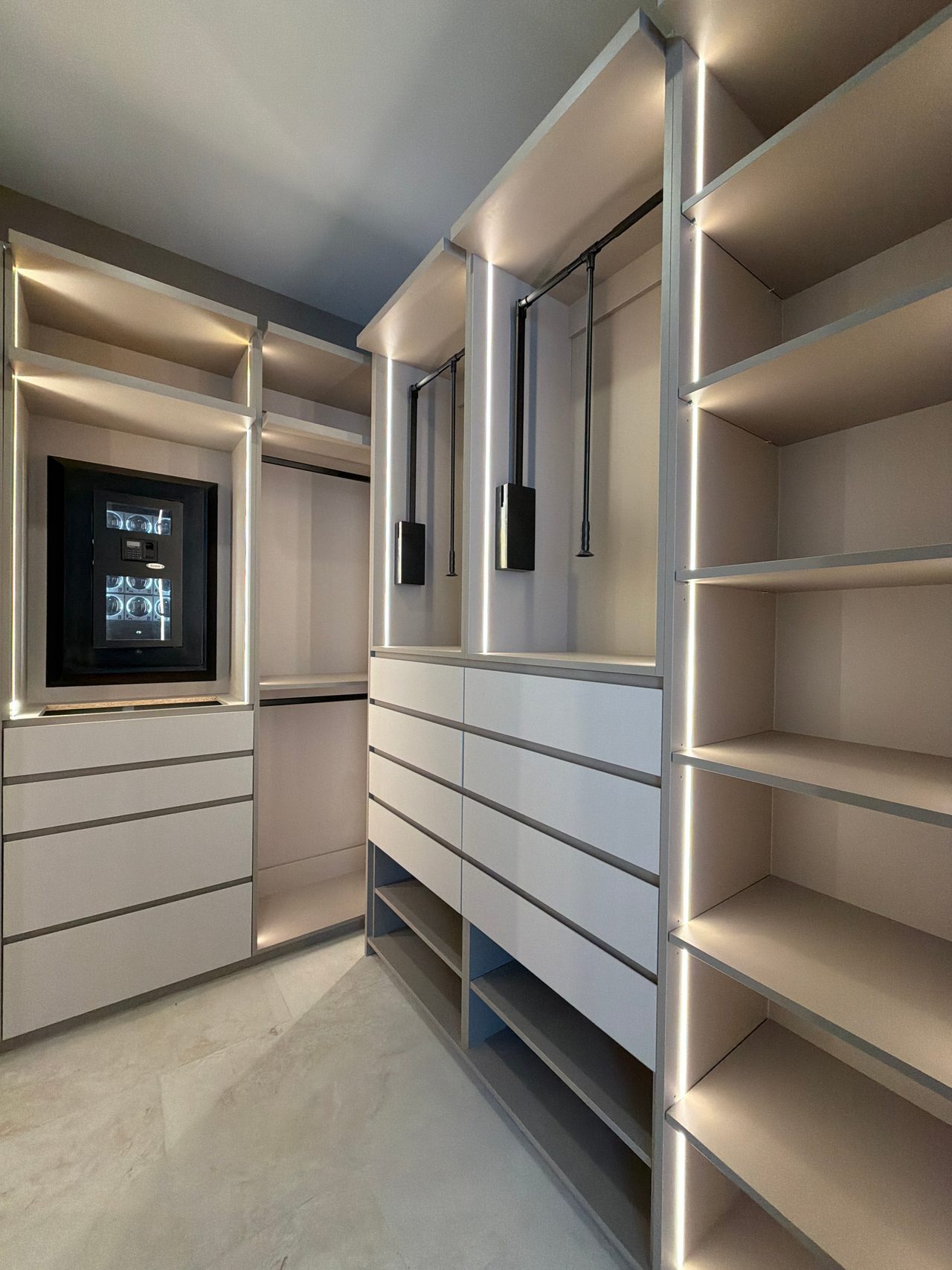 Walk-in closet aisle with pull-down hanging rods, white drawer banks, LED-lit open shelving and a lit glass-front display case in the wall