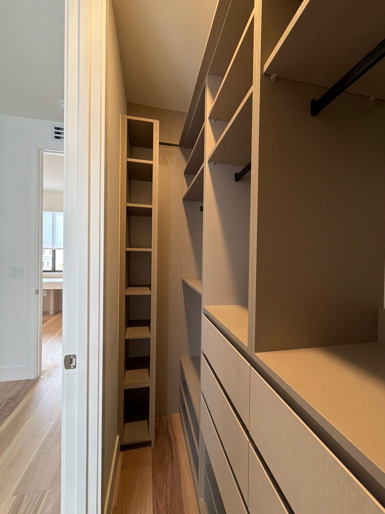 Walk-in closet in beige with a tall narrow cubby tower, open shelves, black hanging rods and a bank of flat-front drawers on a wood-grain floor