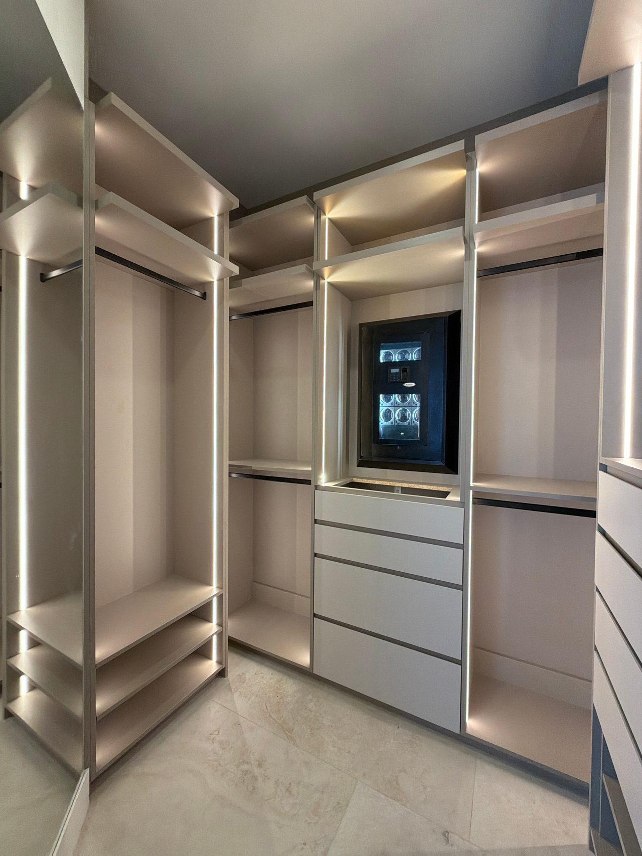 Walk-in closet corner with LED-lit hanging sections, a white drawer stack and an illuminated glass-front display case, stone-look tile floor