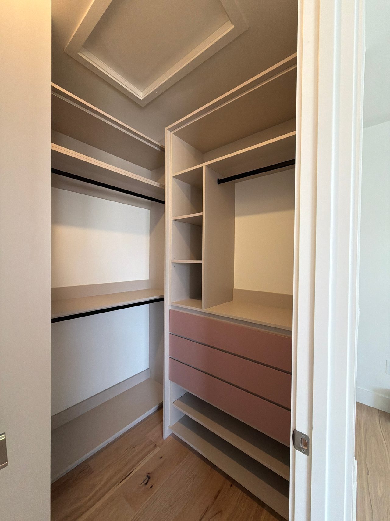 Walk-in closet corner with pink drawer fronts, greige open shelves, black hanging rods and a recessed tray ceiling above a wood-grain floor