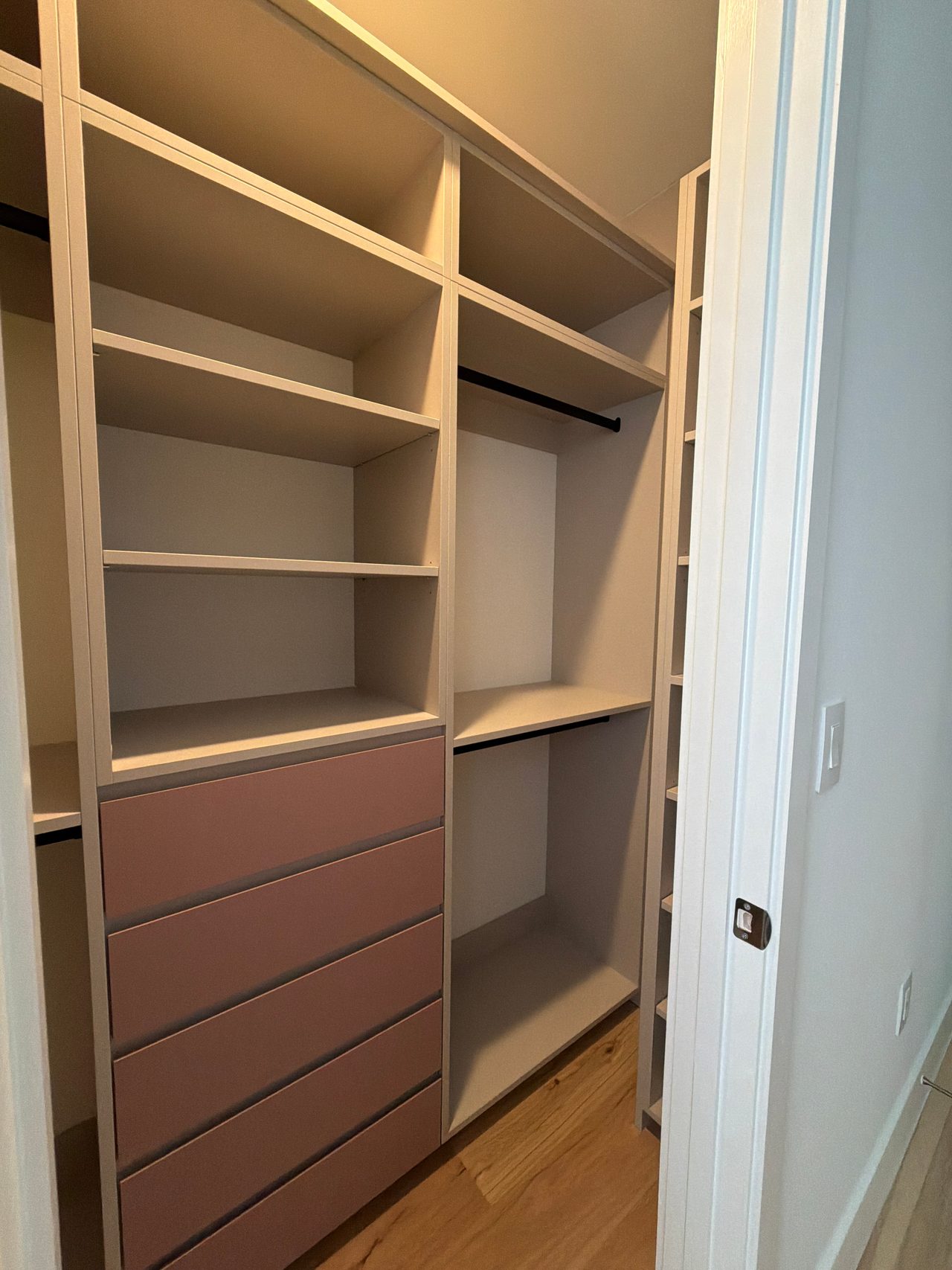 Walk-in closet with cream open shelving, black hanging rods and a five-drawer stack in mauve-pink at left, next to a narrow cubby column