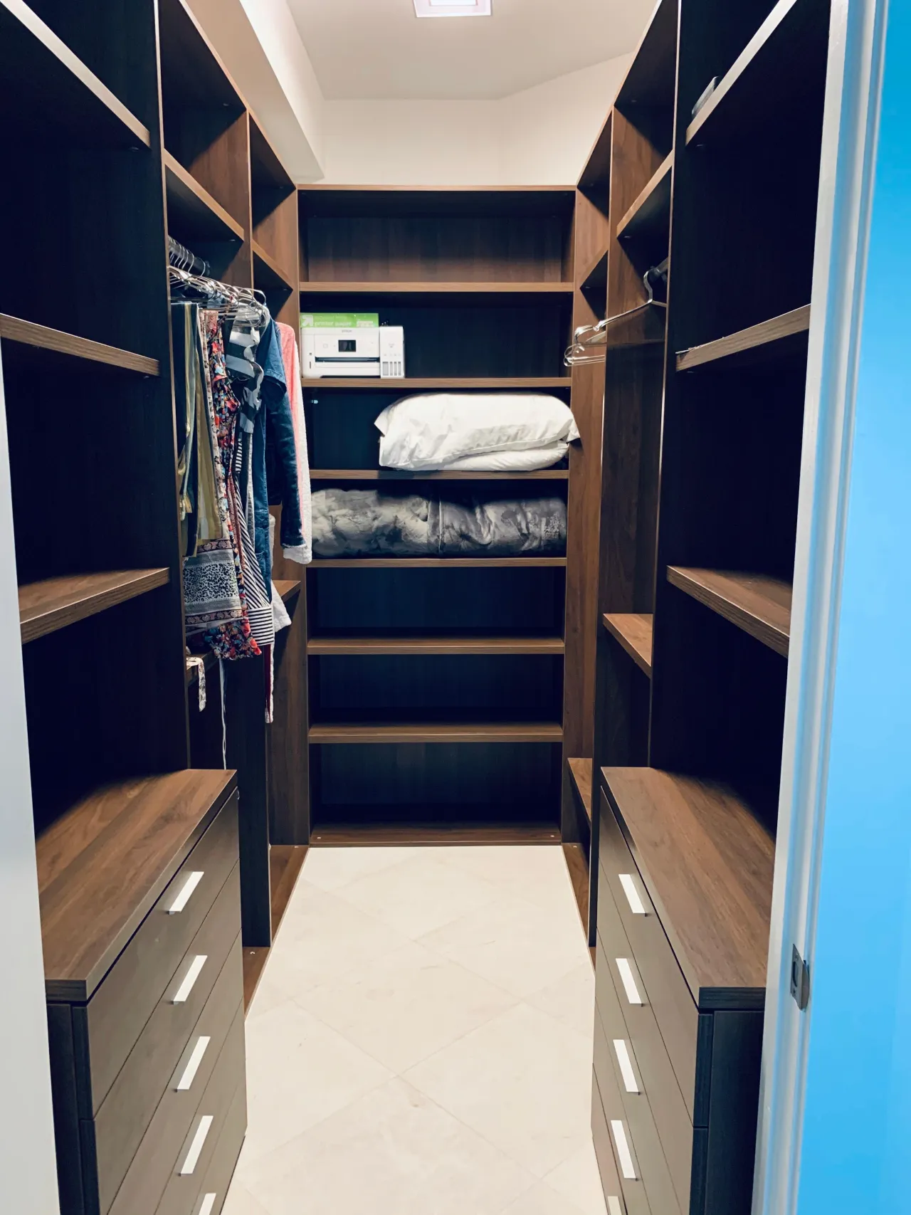 Narrow walk-in closet in dark wood-grain with clothes on a rod at left, open corner shelving holding folded bedding, and drawer stacks with slim pulls