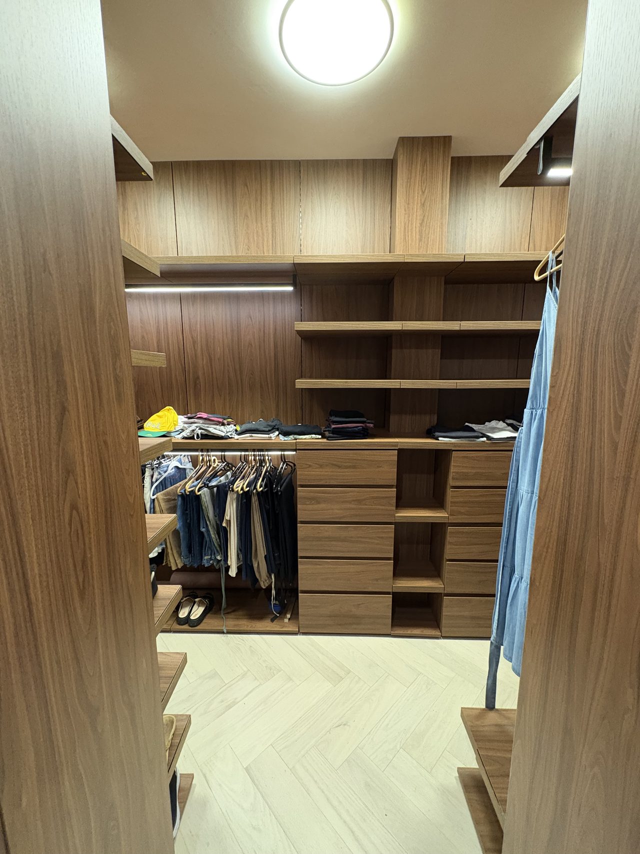 Walk-in closet in dark wood-grain melamine with a central drawer bank, open shelves, folded clothes on the counter, and hanging trousers