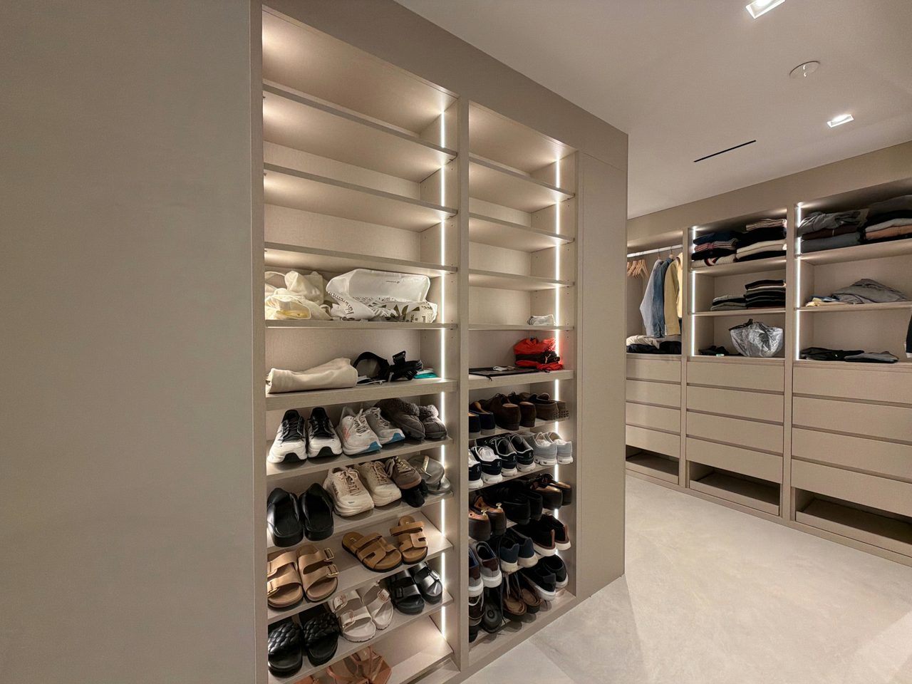 Walk-in closet with gray melamine open shoe shelving lit by vertical LED strips, a hanging section, and drawer banks under lit upper shelves
