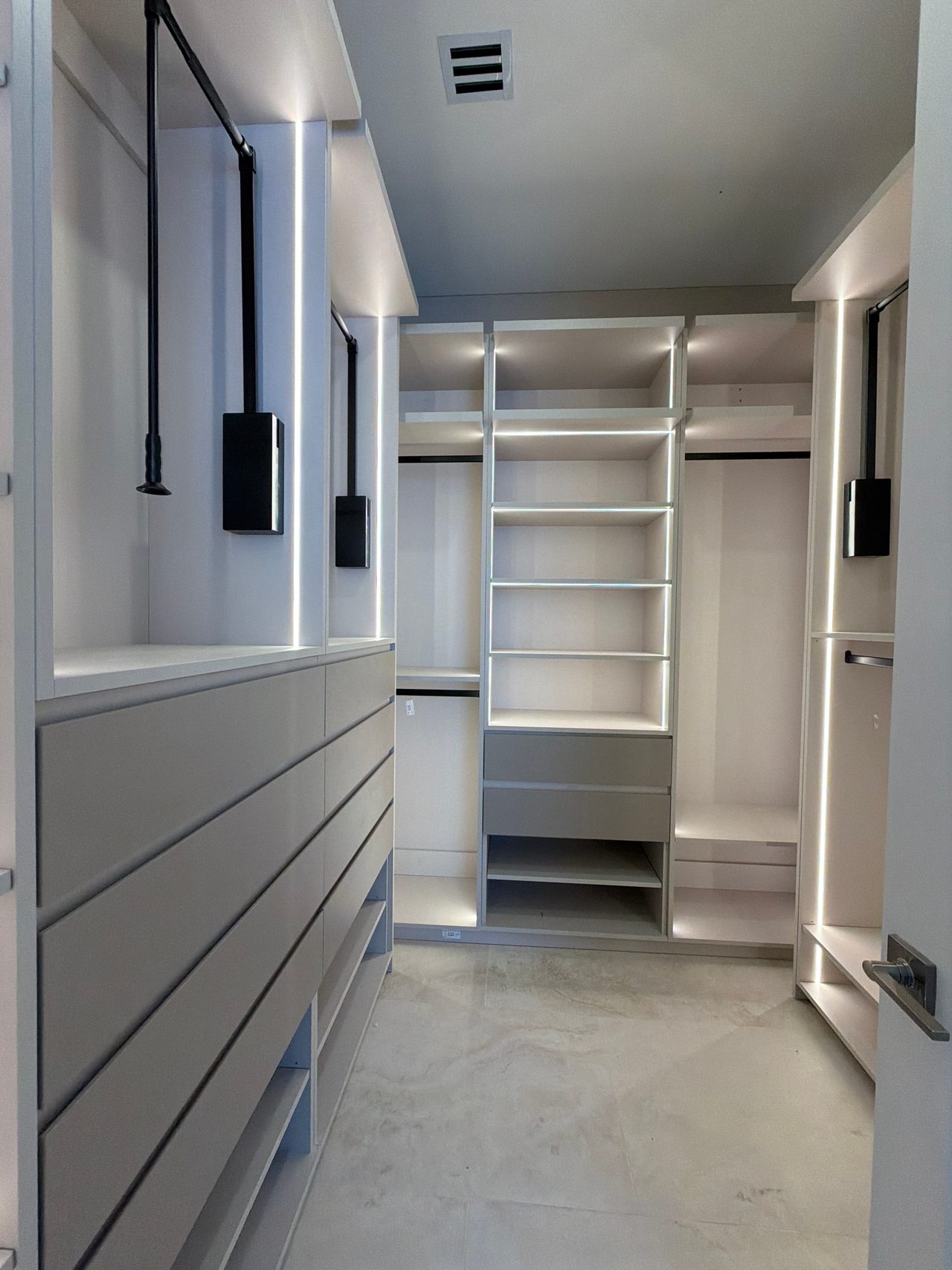 Walk-in closet with pull-down hanging rods, a long white drawer bank at left, a central LED-lit shelf tower and a stone-look tile floor