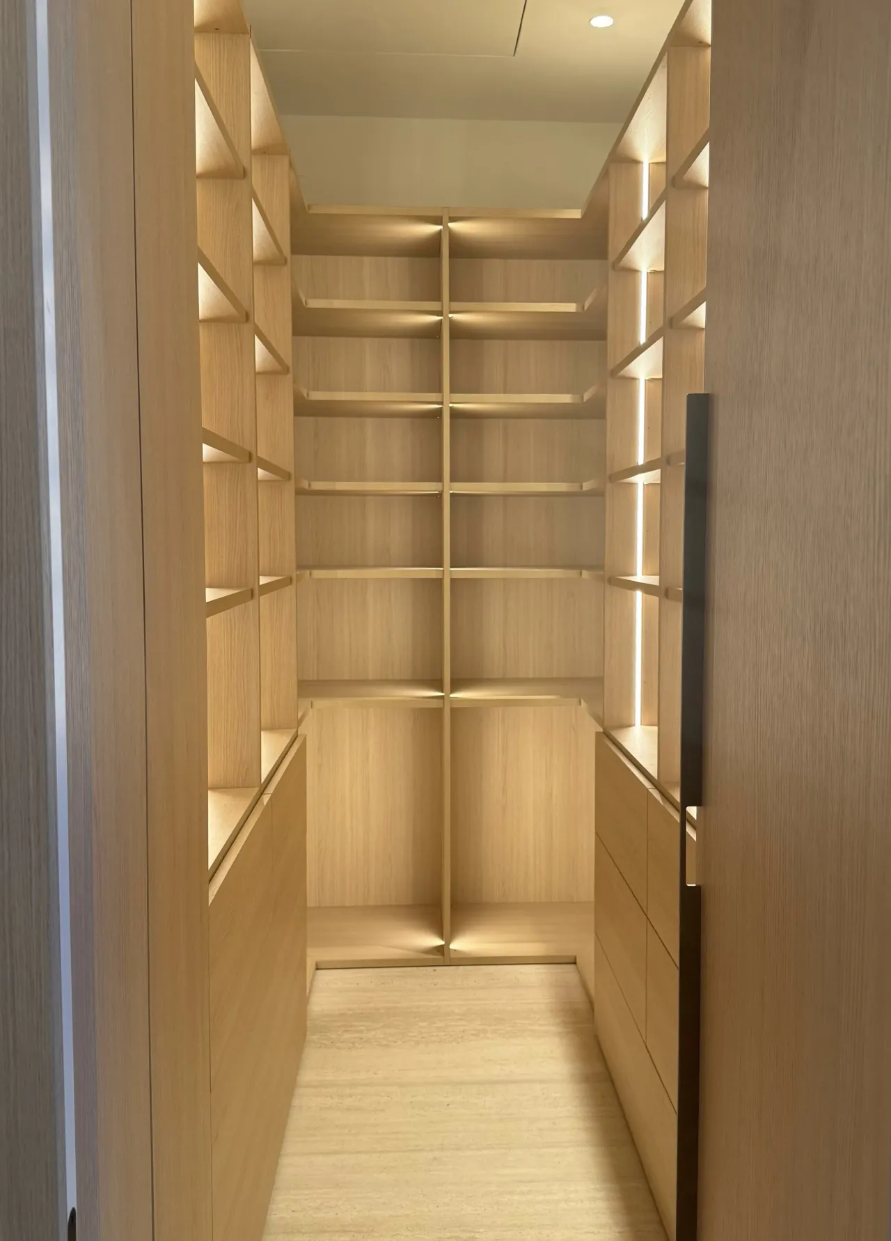 Walk-in closet with tall open shelving on both sides and drawer stacks below, LED edge lighting throughout, light stone floor