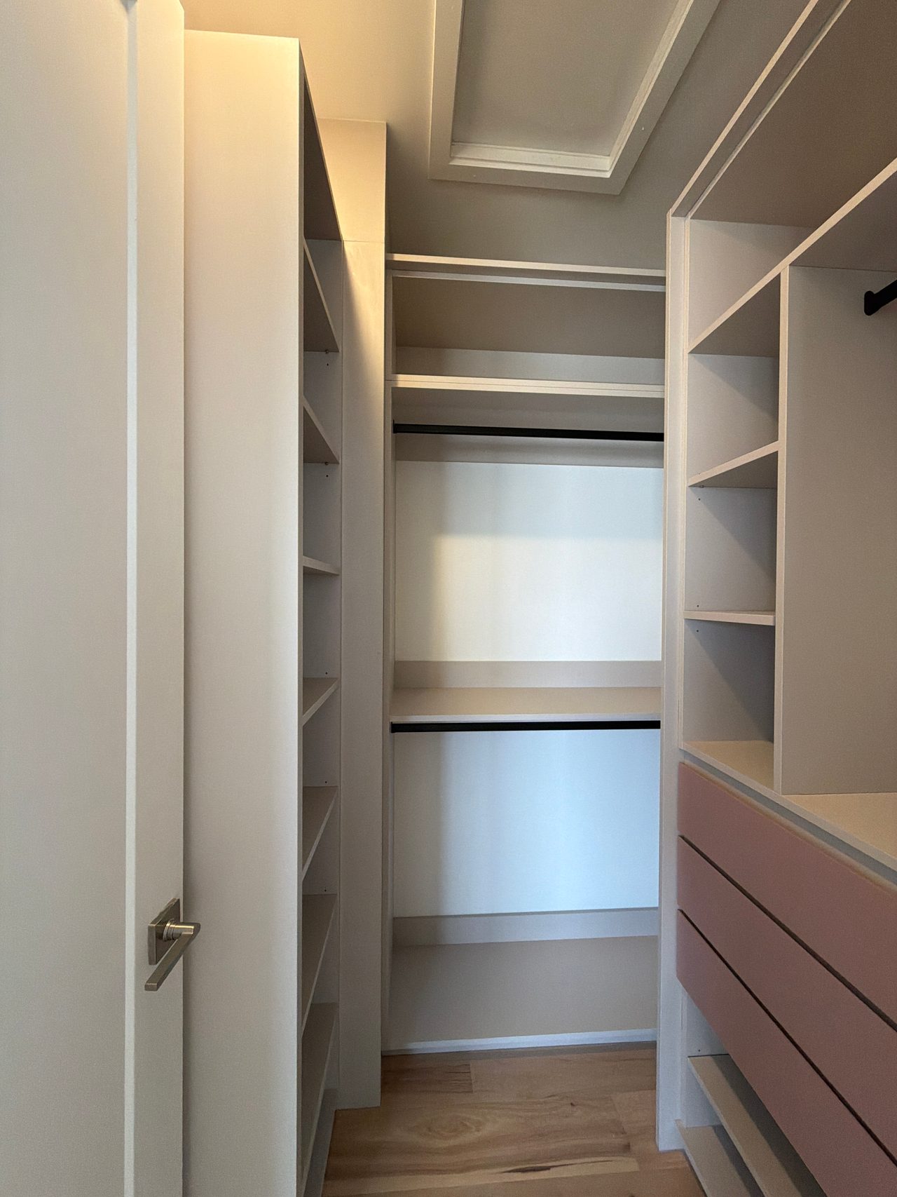 Narrow walk-in closet with a tall shelf tower at left, two stacked black hanging rods on the back wall and pink drawer fronts at right