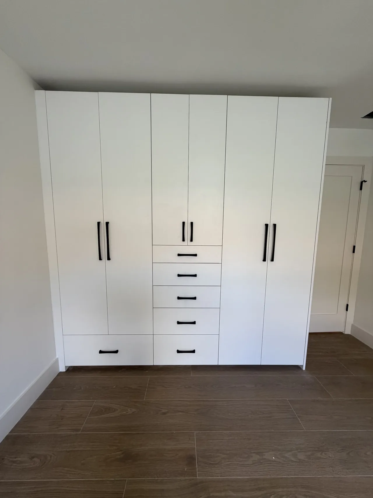 White floor-to-ceiling wardrobe, six doors with black bar pulls, one center column of five drawers and a wide drawer at lower left, dark wood-look floor