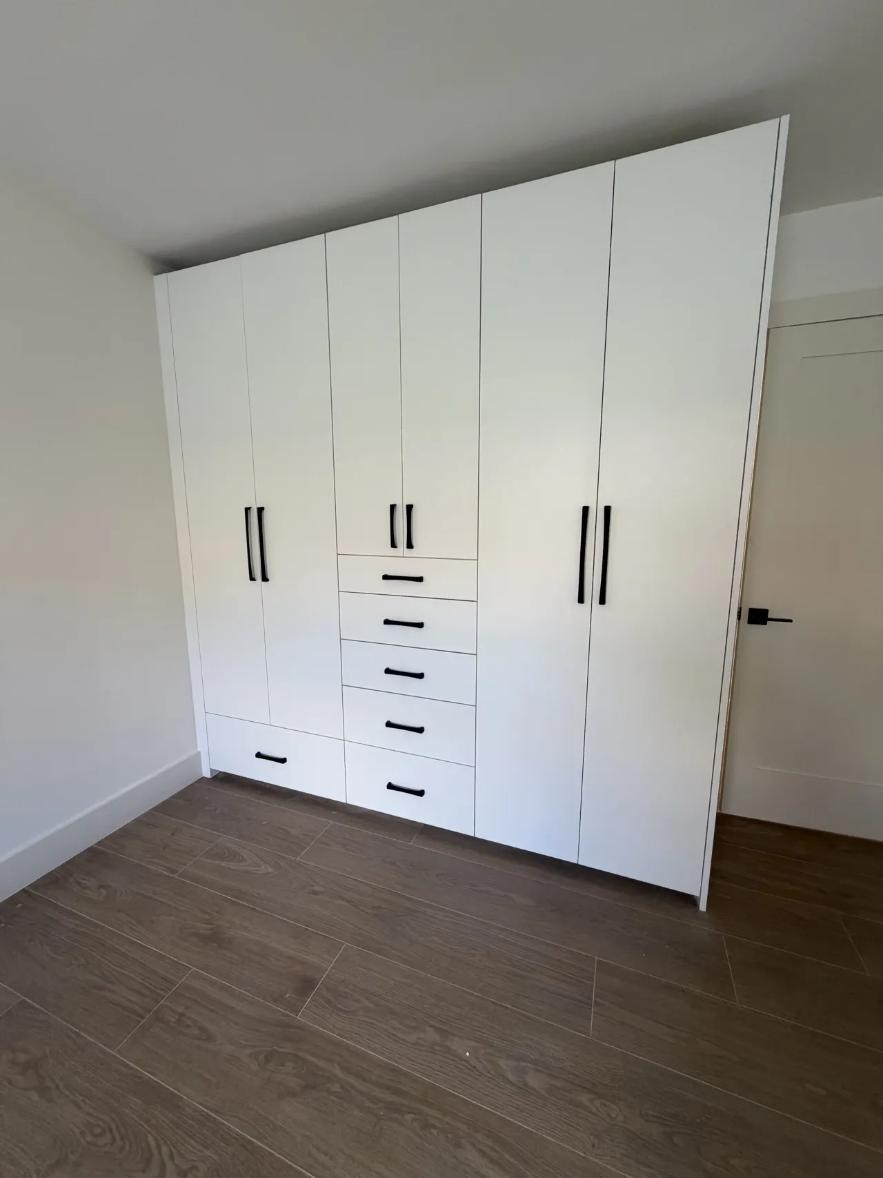 White floor-to-ceiling wardrobe with six doors and a center column of drawers with black pulls, on dark wood-look flooring