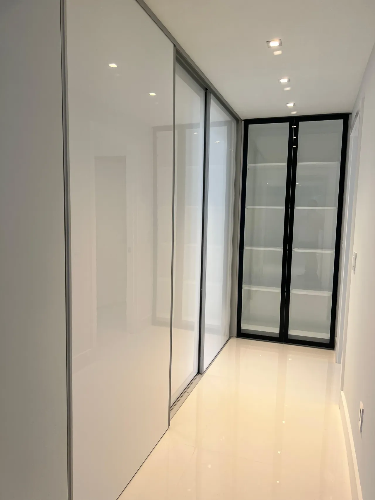 White glossy sliding closet doors beside a black-frame glass display cabinet with visible shelves, glossy light stone floor hallway